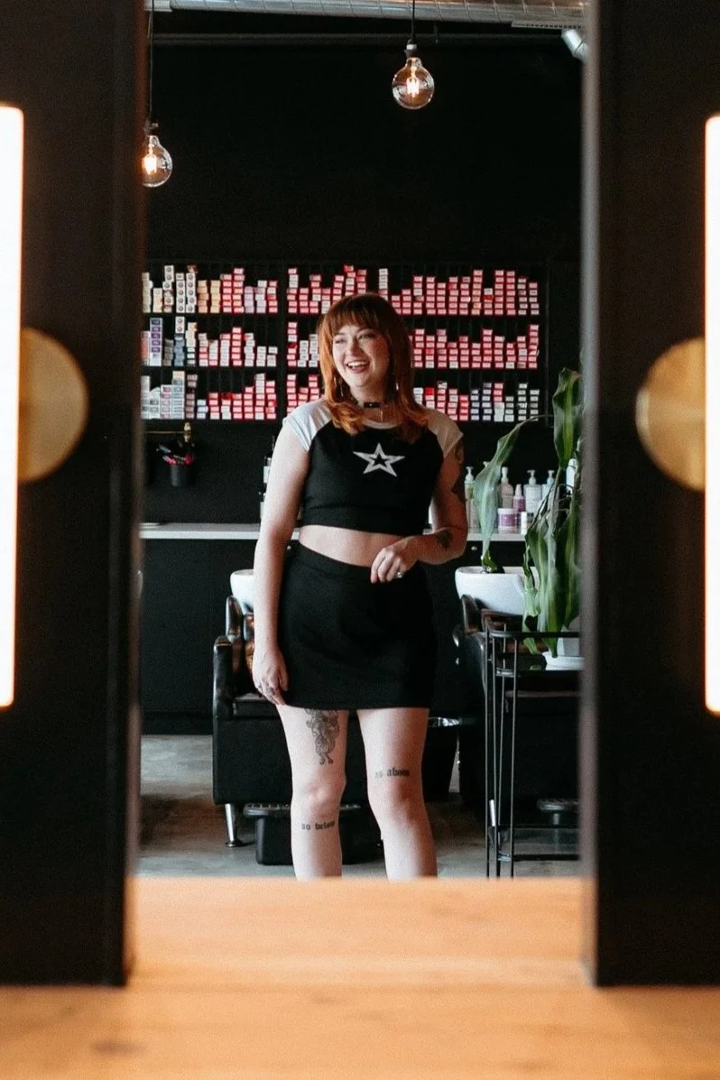 Taylor, with red hair and tattoos smiling at herself in a mirror inside a modern salon, with shelves of hair color and products behind her.