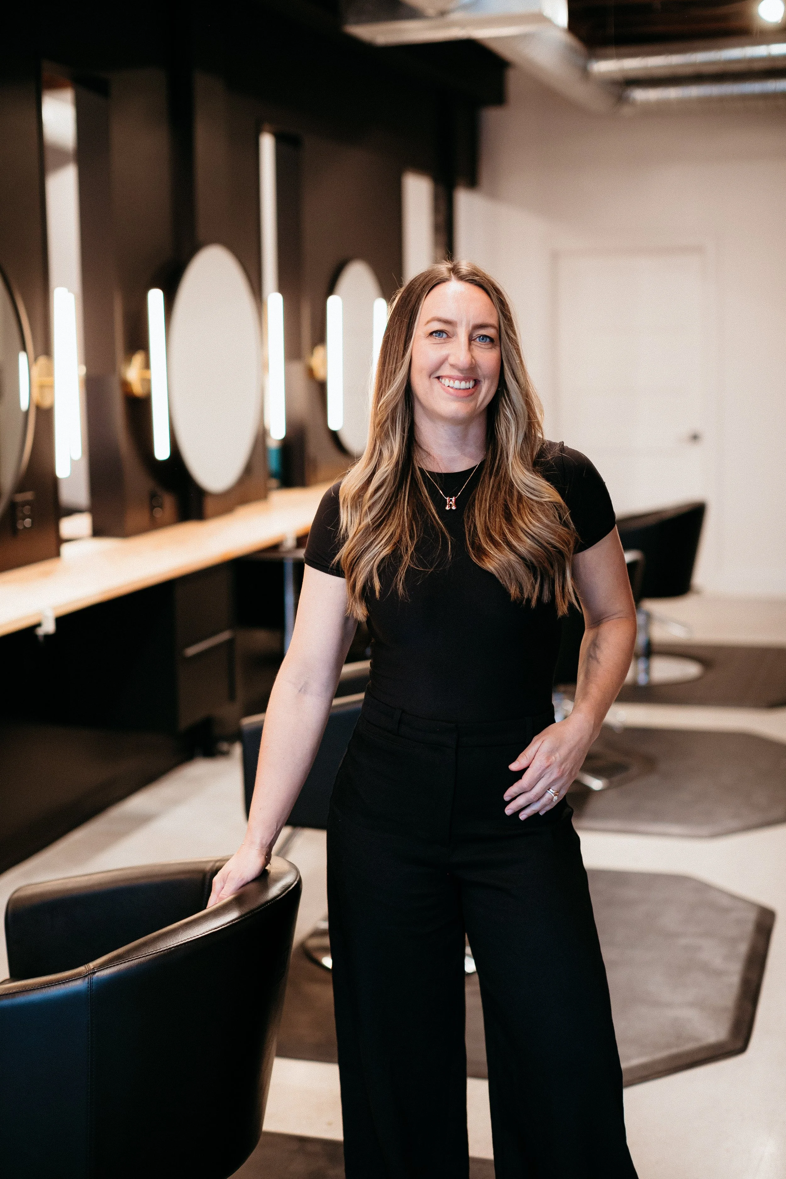 Heather with long wavy hair wearing a black dress standing in a modern salon or barber shop, with hair styling mirrors and chairs in the background.