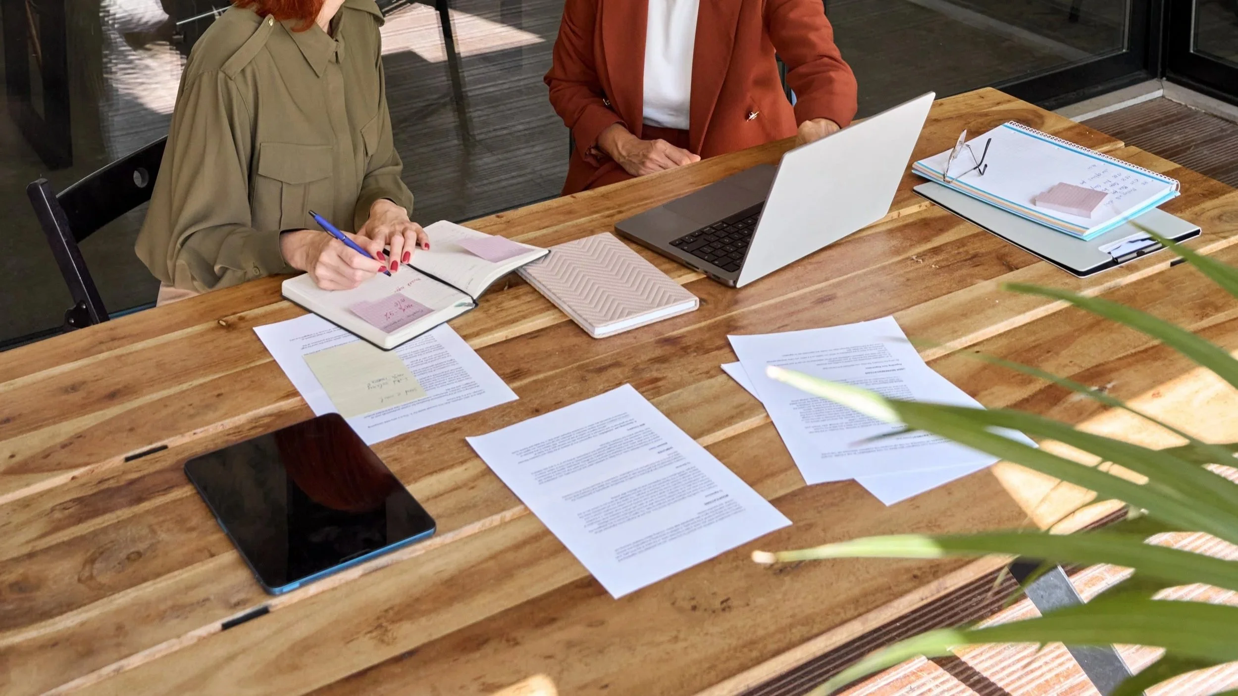 Two women working at a wooden table filled with paperwork, notebooks, a laptop, glasses, and a tablet.