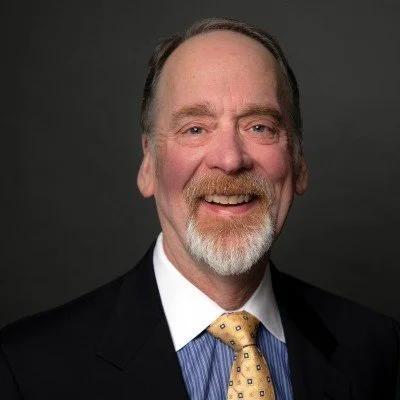 Headshot of a man wearing a navy blazer, blue dress shirt, and yellow tie