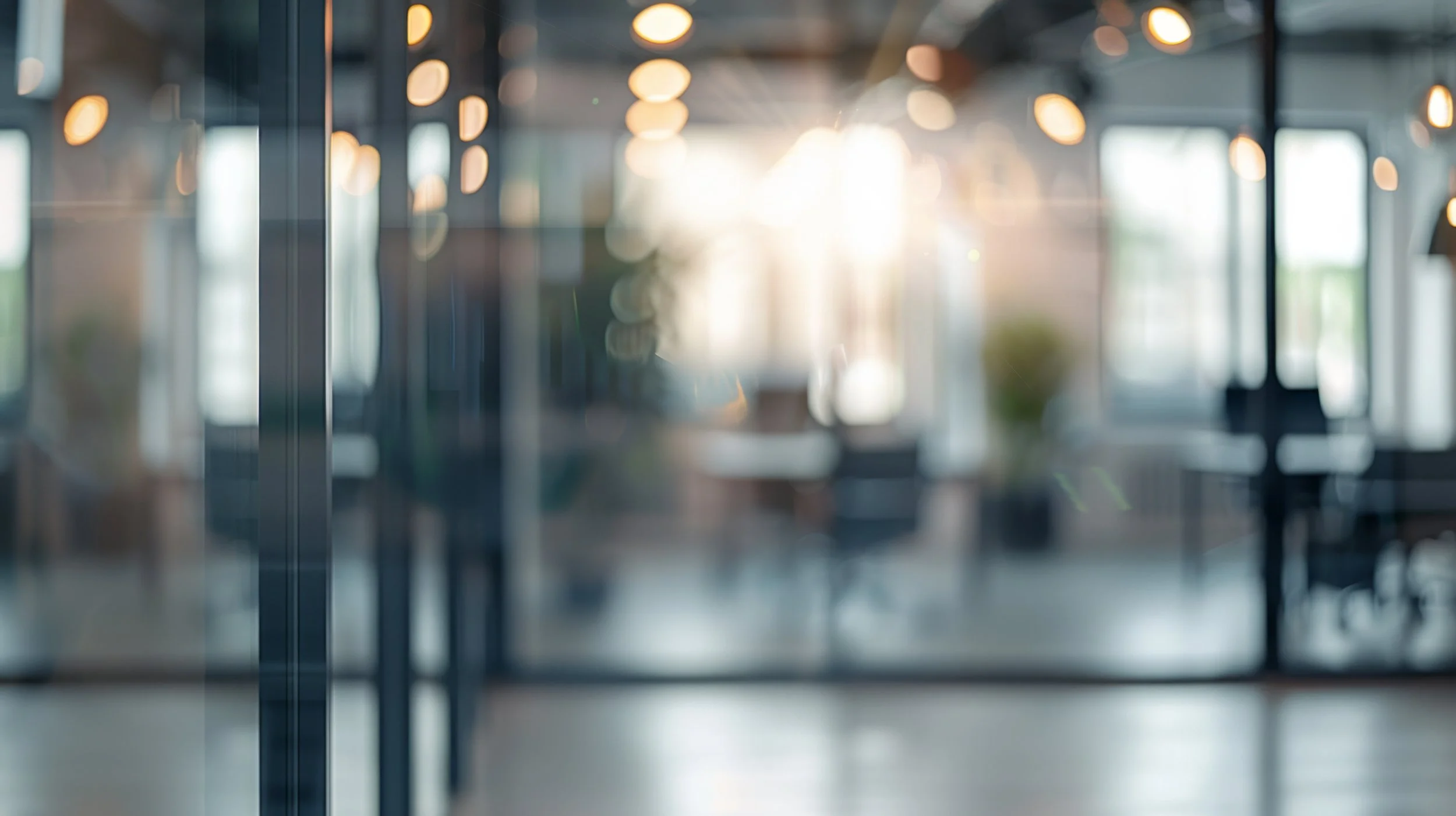 Blurry view of an indoor space with hanging lights and large windows, sunlight streaming through.