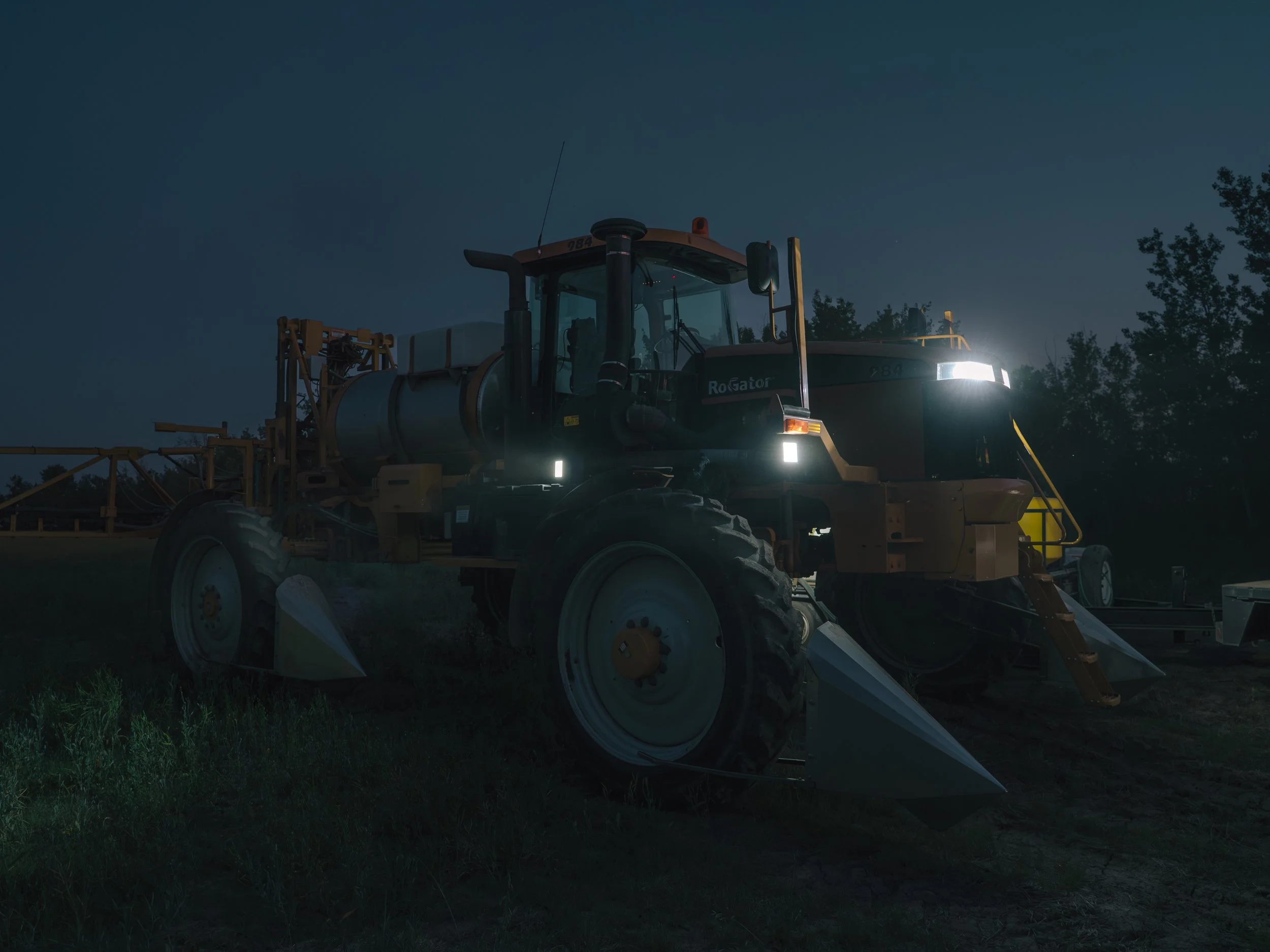 A large agricultural sprayer tractor at night with headlights on, parked on a field with trees in the background.