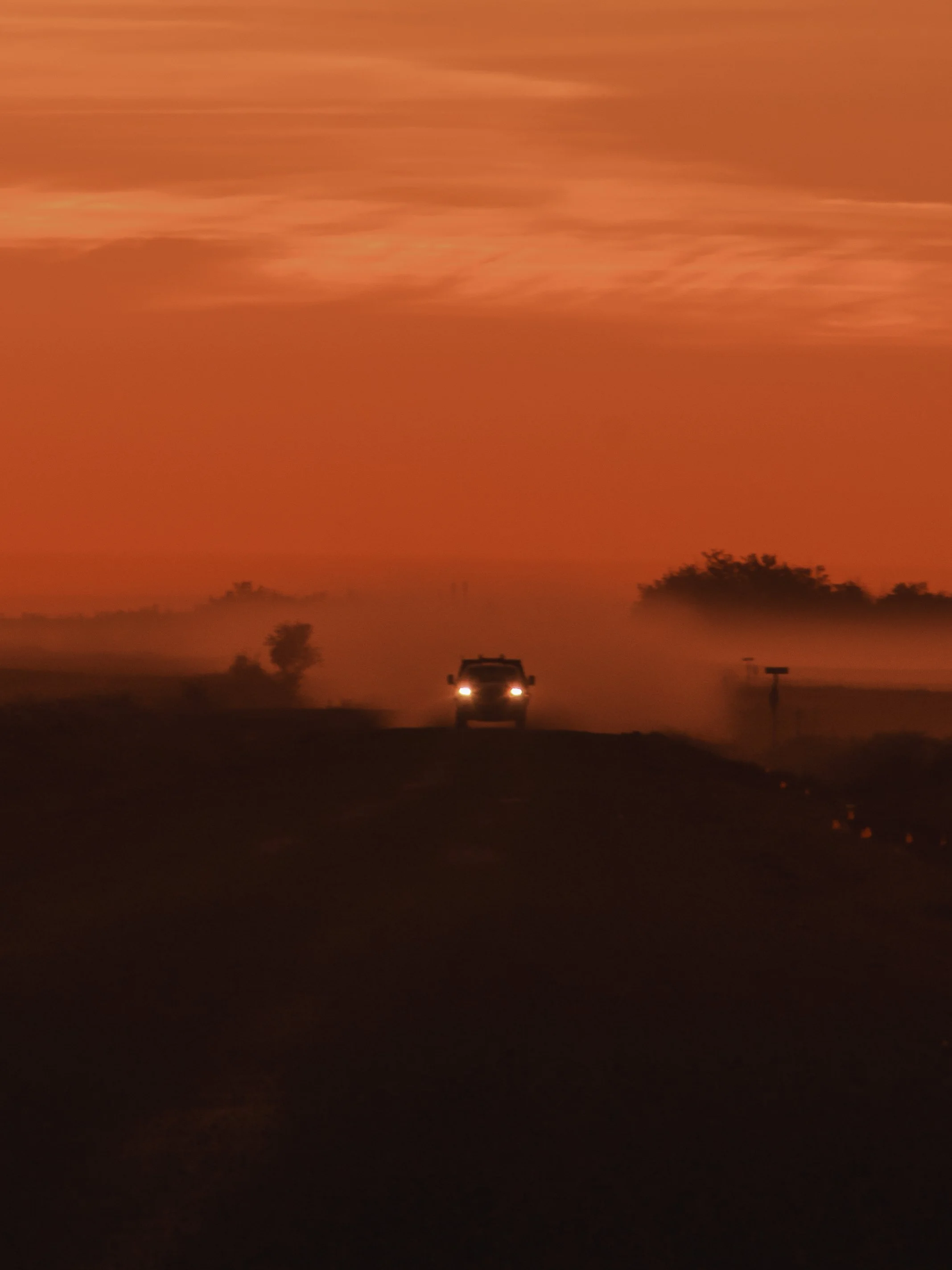 A vehicle driving on a dark road at dusk or dawn with its headlights on, a dust trail behind, against an orange and pink sky with some clouds and distant trees.