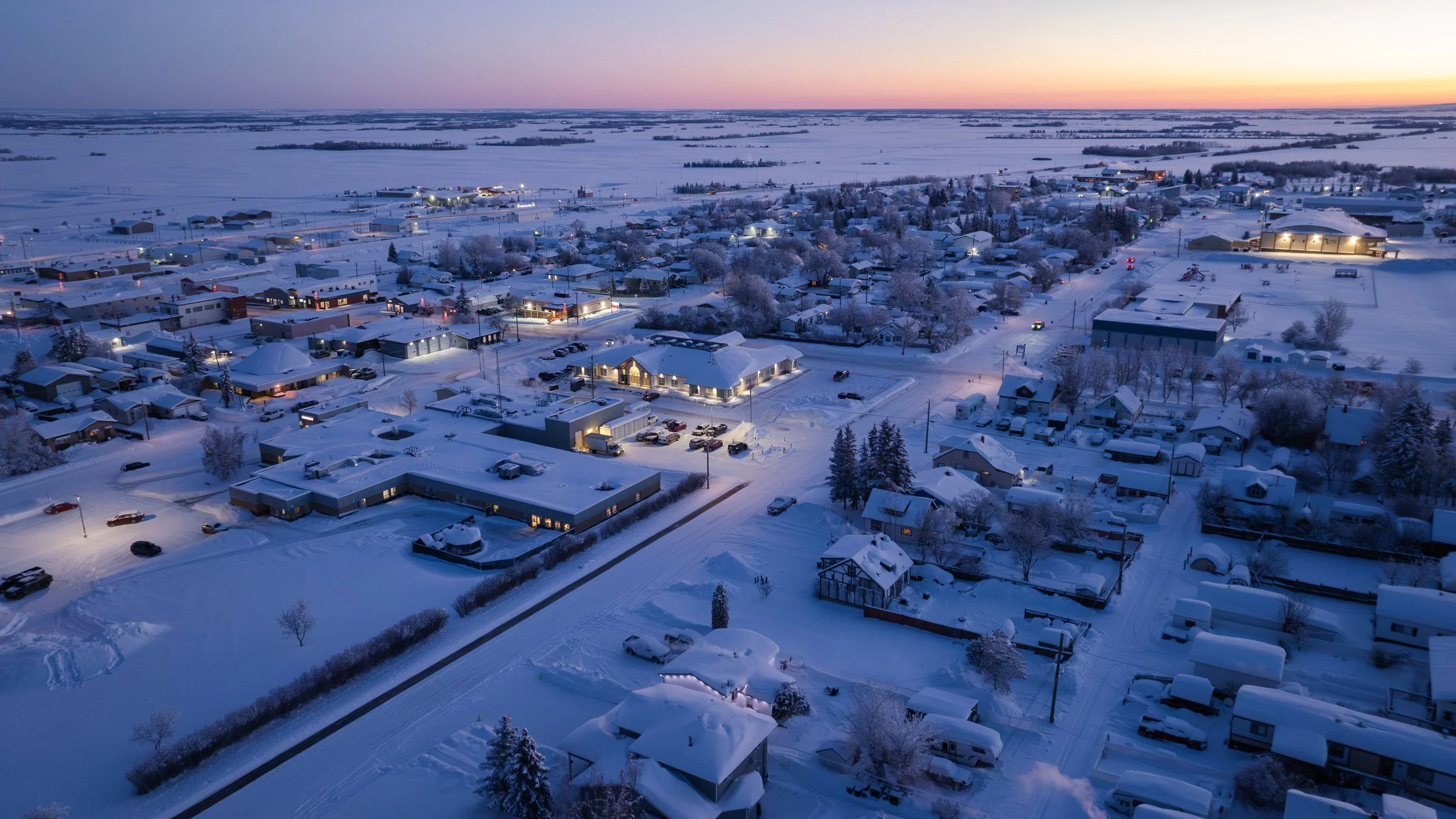 Aerial view of a snow-covered suburban neighborhood at dusk with houses, roads, and trees dusted with snow, and streetlights illuminating the area.
