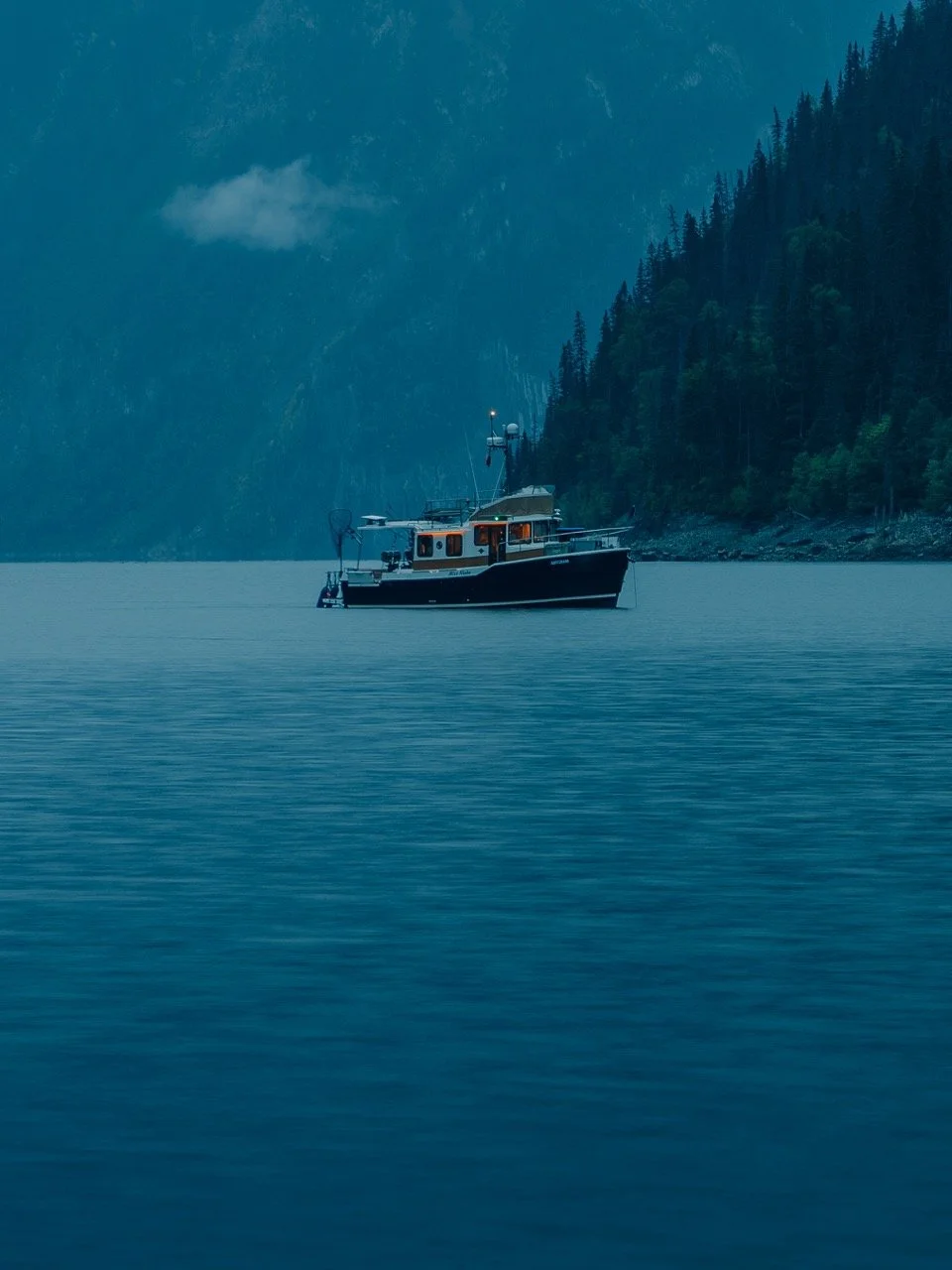 A boat floating on calm water with forested mountains in the background under overcast sky.