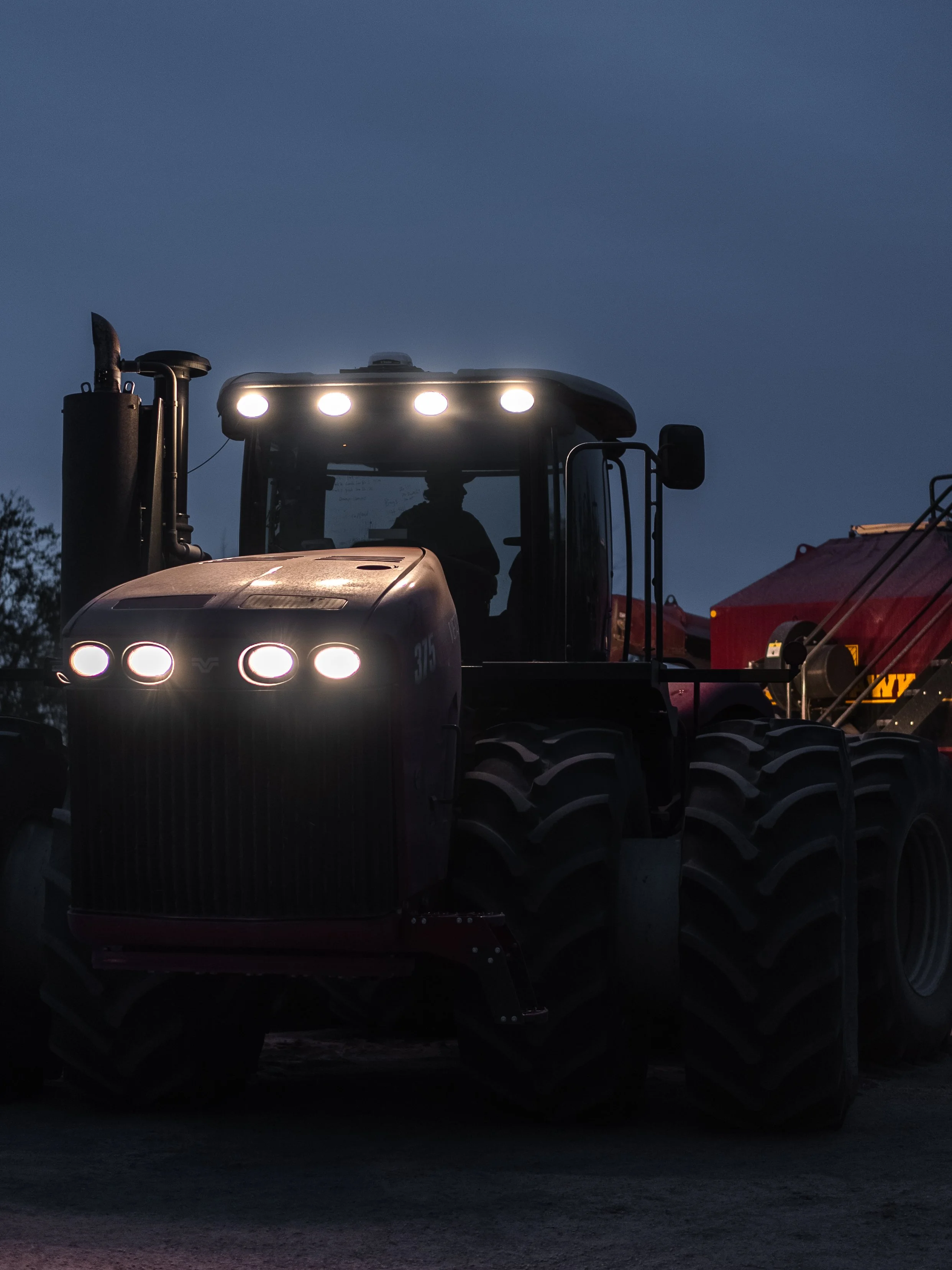 Silhouette of a large tractor with bright headlights on, set against a dusky sky.
