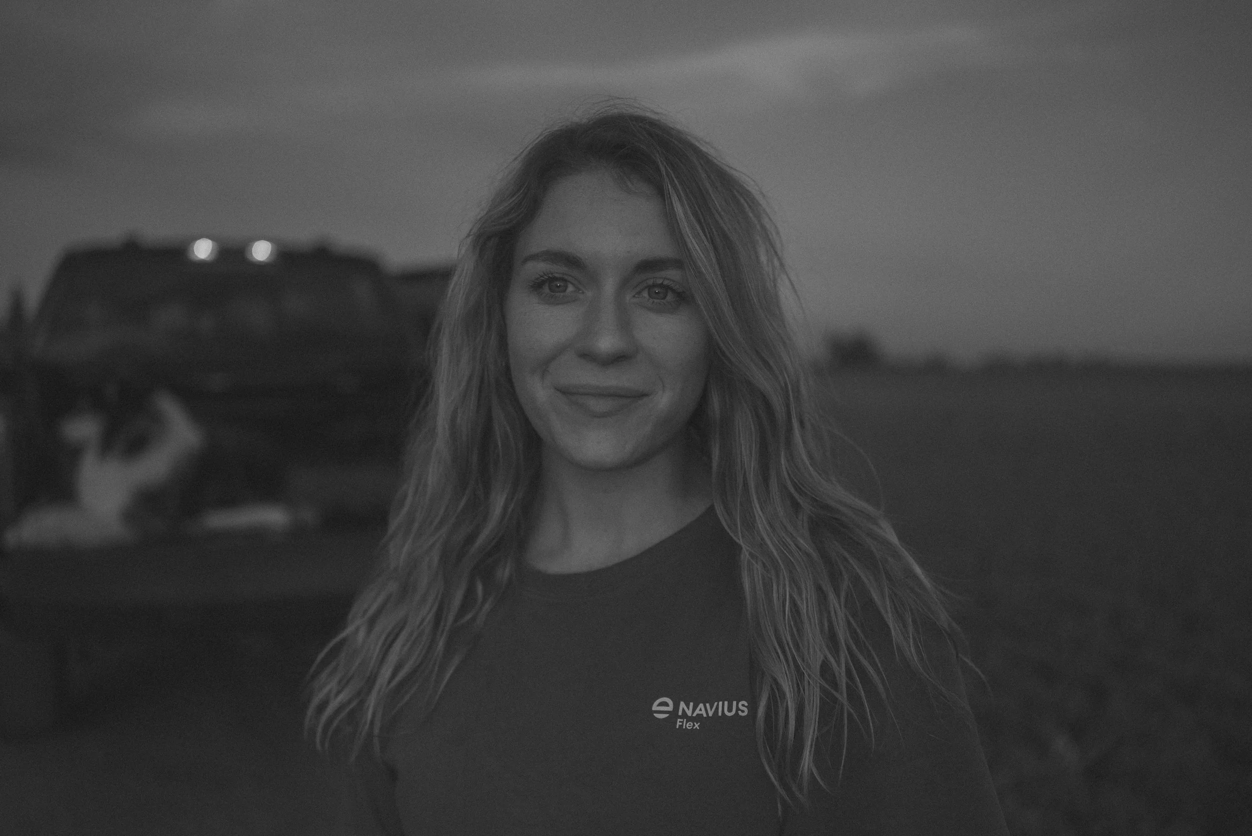 A woman with long, wavy hair standing outdoors during dusk, smiling slightly, wearing a dark NAVIUS Flex shirt.