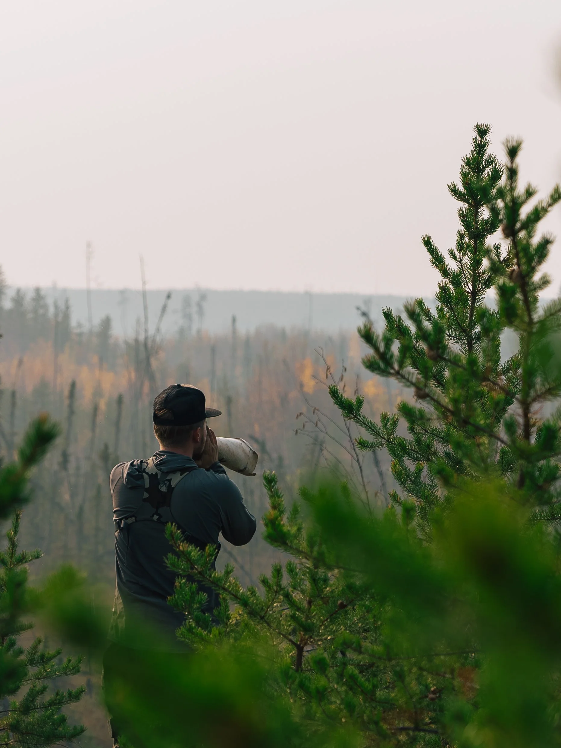 A person in outdoor gear looks through binoculars in a forested area.