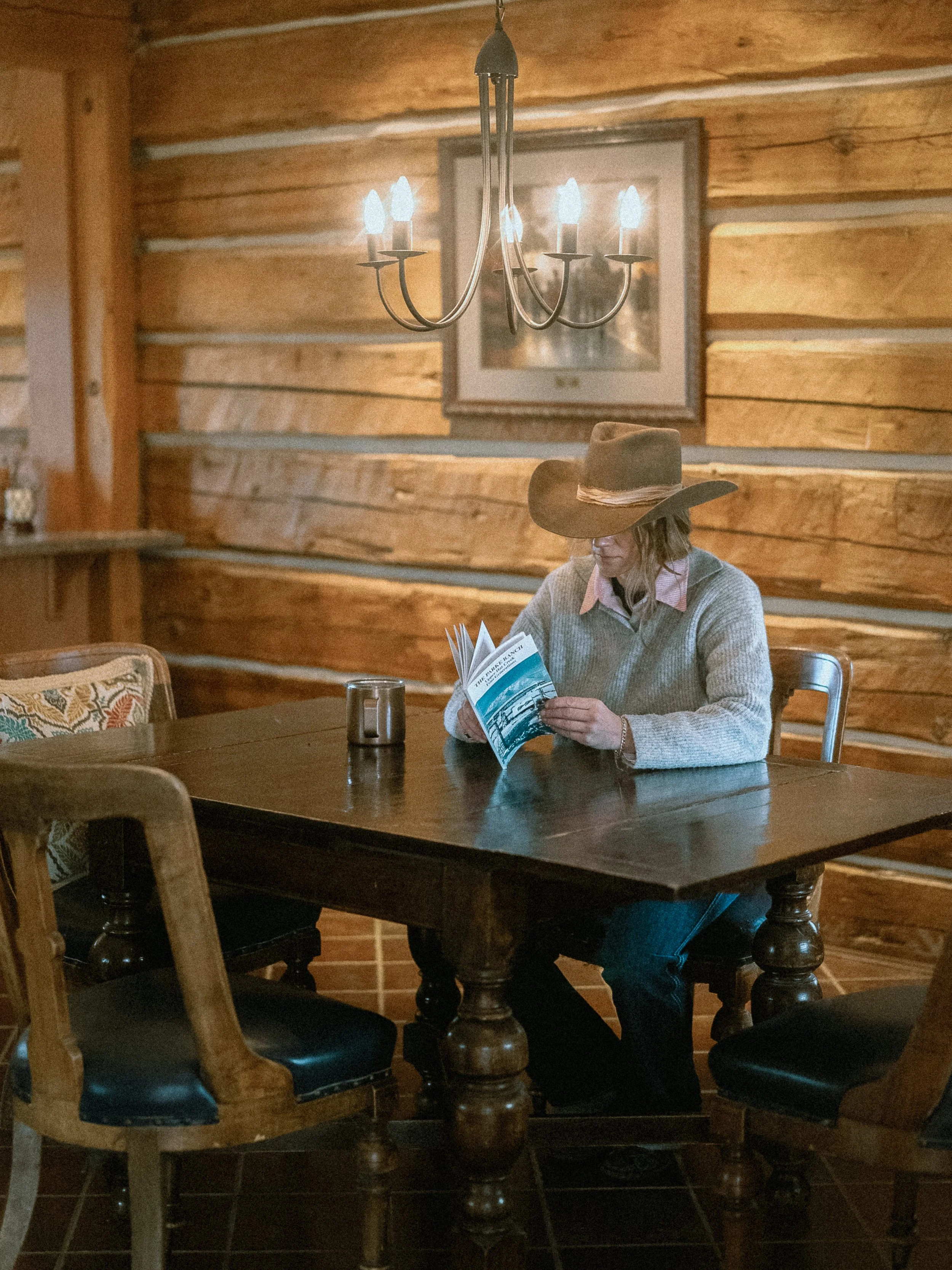 A woman wearing a wide-brimmed hat and sweater, sitting at a wooden table in a rustic cabin, reading a brochure or magazine. The cabin has wooden walls and a chandelier hanging above.