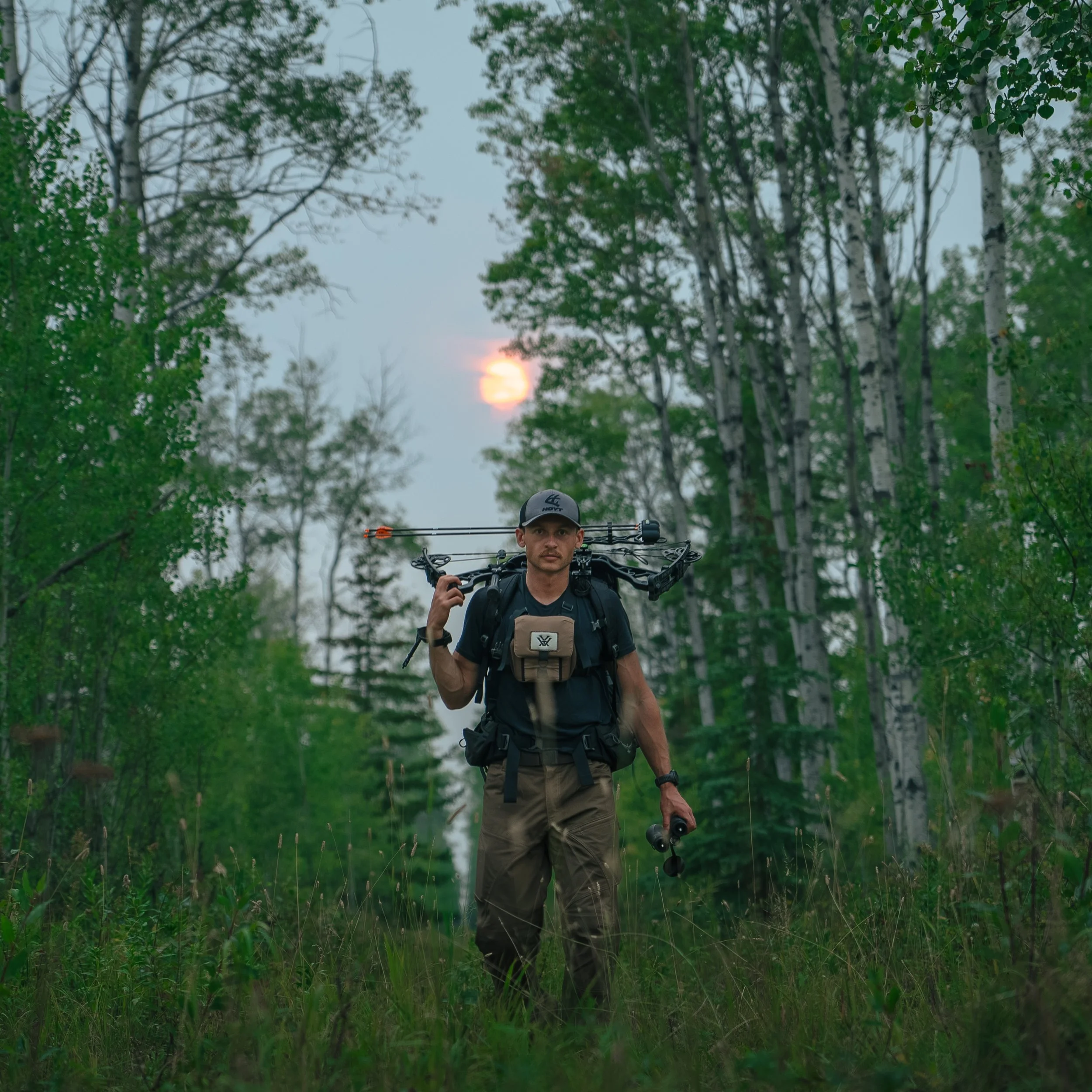 Man hiking in a forest at dusk, carrying a bow and a backpack, with tall trees and the moon visible in the cloudy sky.