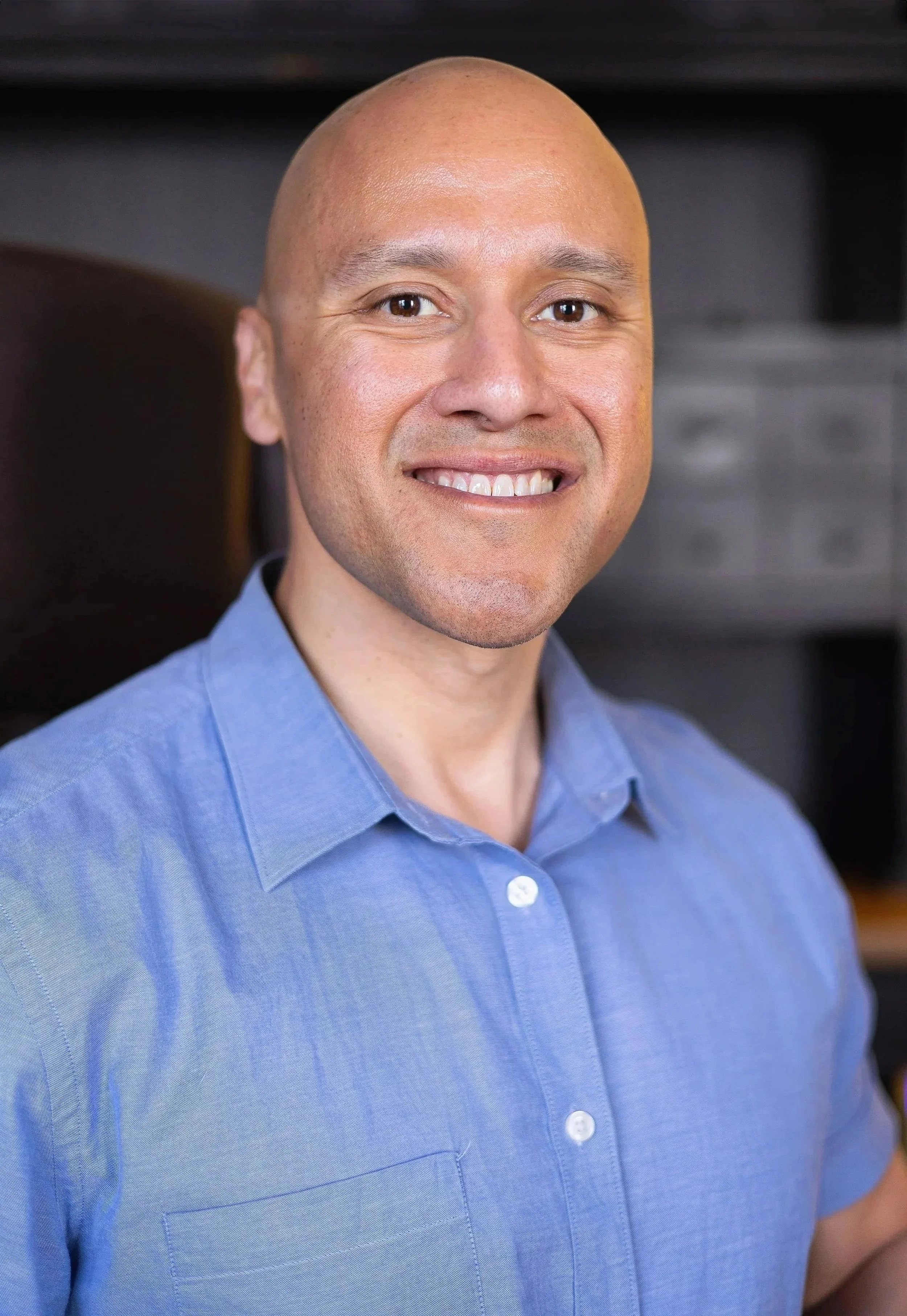 A smiling man with a shaved head wearing a blue collared shirt, standing indoors with dark furniture in the background.