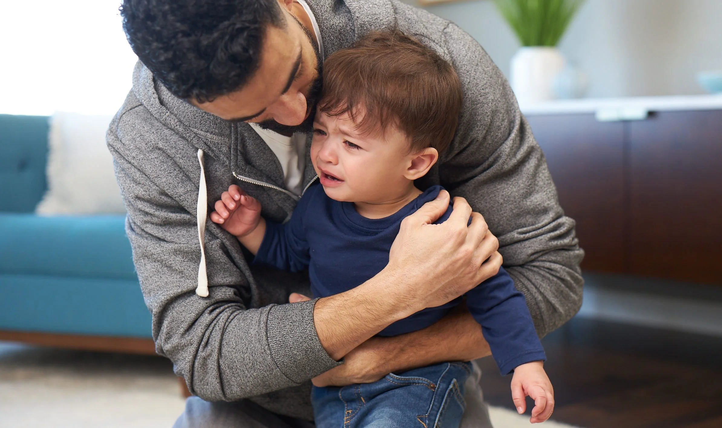 A man in a gray hoodie comforting a crying young boy in a blue shirt indoors.