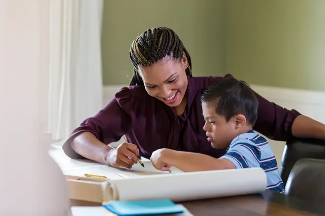 A woman and a young boy sitting at a table, working together on a large piece of paper with coloring supplies nearby.