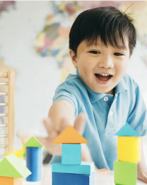 Smiling young boy playing with colorful geometric building blocks in a room with a blurred mural background.