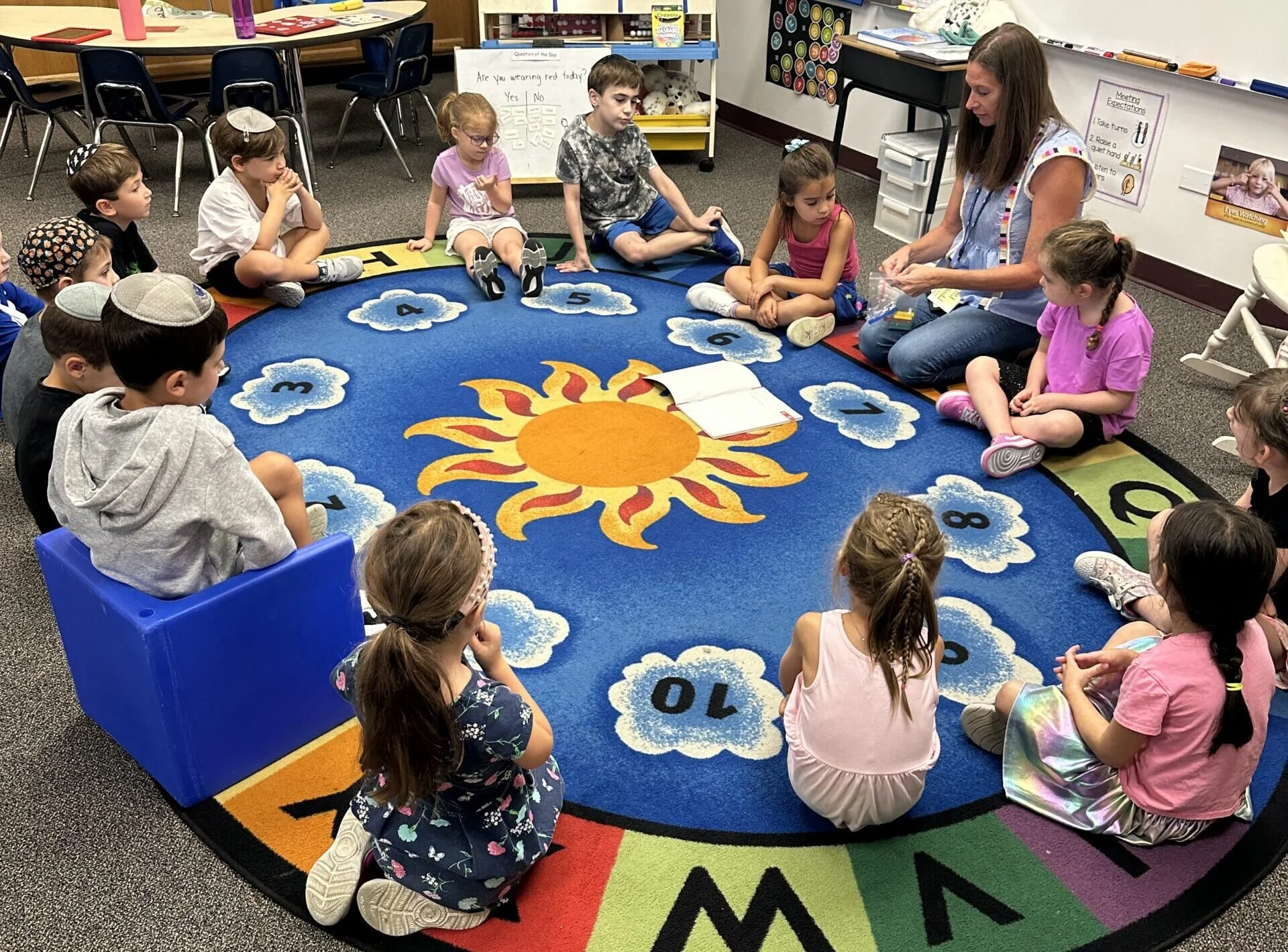 A teacher sitting on the floor with children sitting around her on a colorful classroom carpet featuring a sun and clouds. The children are attentively listening during an activity, with some appearing focused and others curious. The classroom has tables, chairs, and educational materials visible in the background.