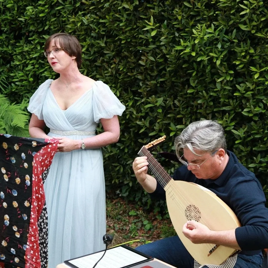 Anastassia Rakitianskaia sings in a blue dress against a tall hedge. Beside her Brian Payne plays the lute.
