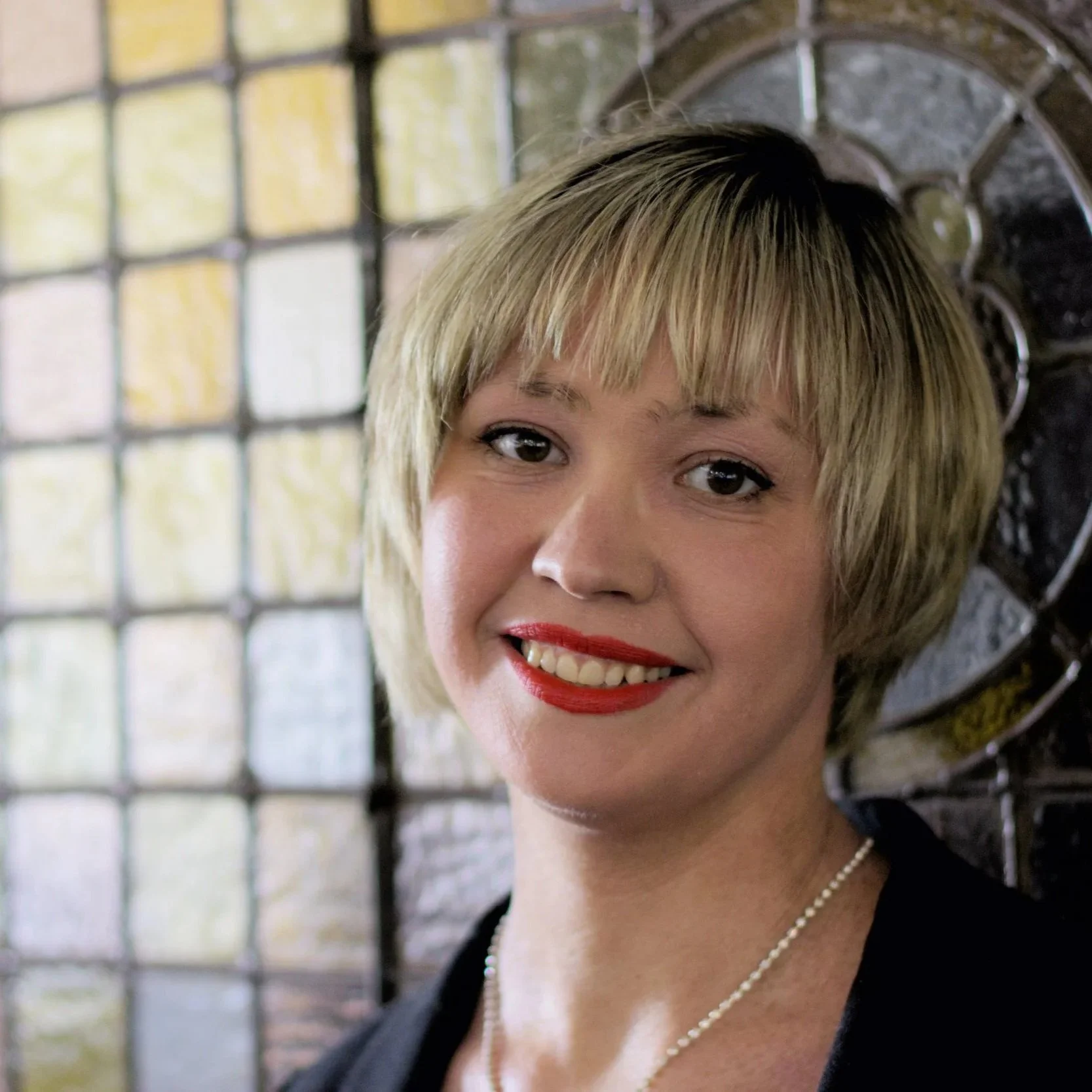 A woman with short blonde hair, red lipstick, wearing a black top and a pearl necklace, standing in front of a stained glass window and a decorative metal wall.