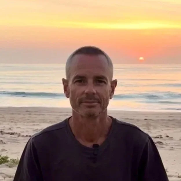 A man with short gray hair and a beard sitting on a beach at sunset, with the ocean and colorful sky in the background.