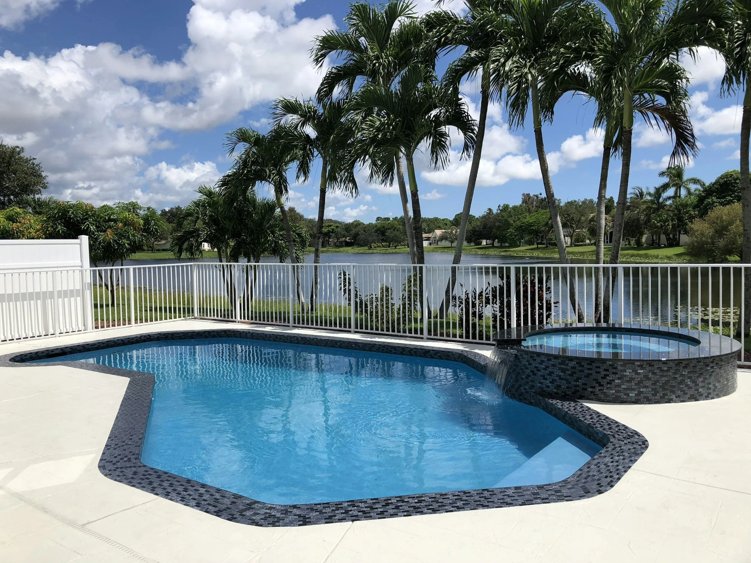 Swimming pool with a black tile border, surrounded by a white fence, overlooking a lake with trees and houses under a partly cloudy sky.