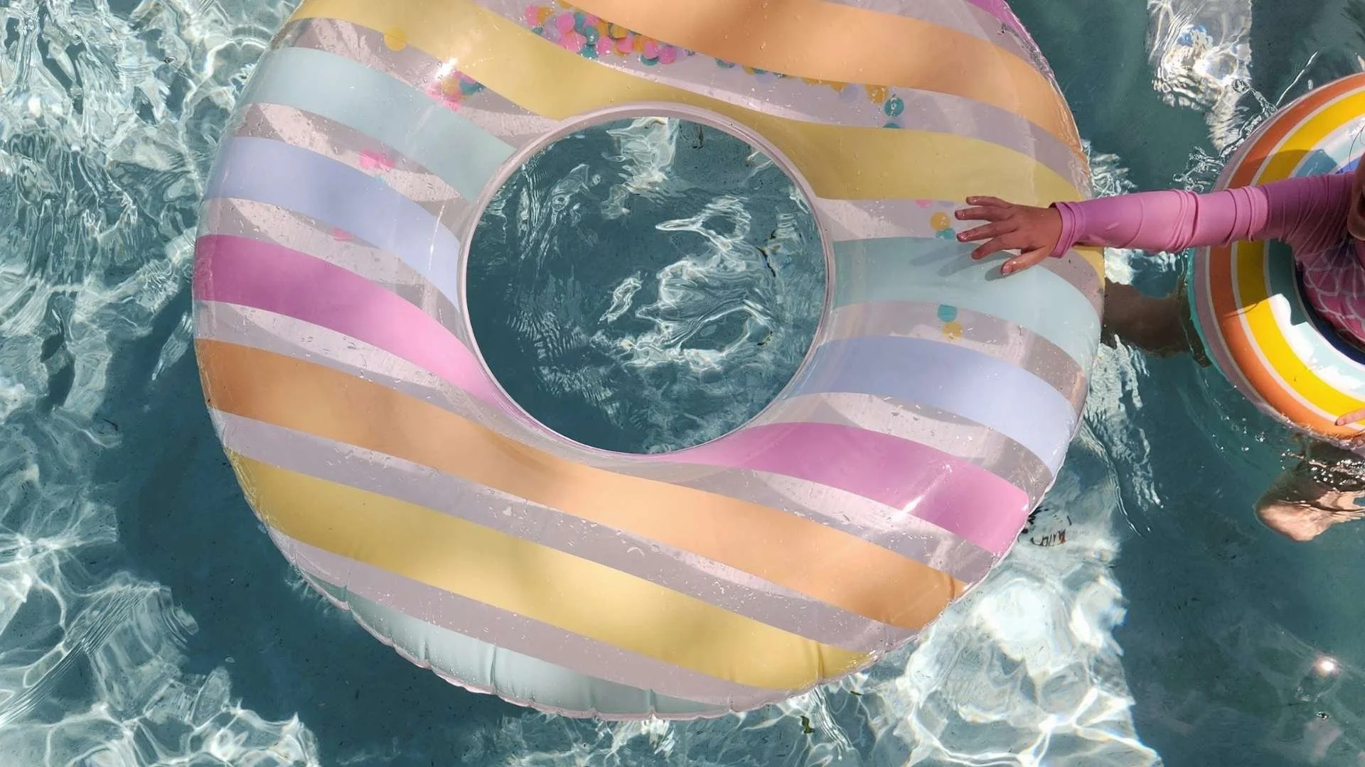 Colorful inflatable pool float with pastel rainbow stripes floating in a pool, with a child's hand reaching out to the float.