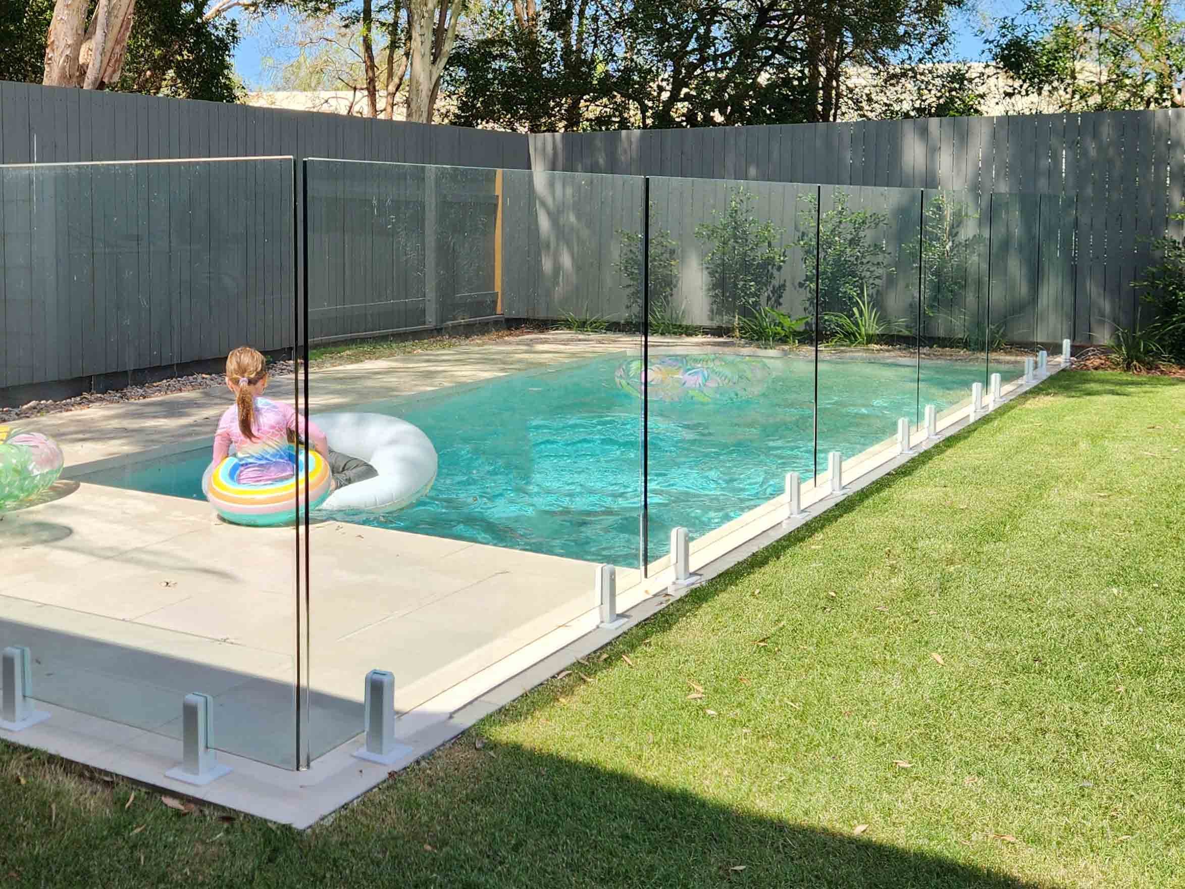 Child sitting on a pool float in a backyard swimming pool, surrounded by a glass safety fence and green grass.