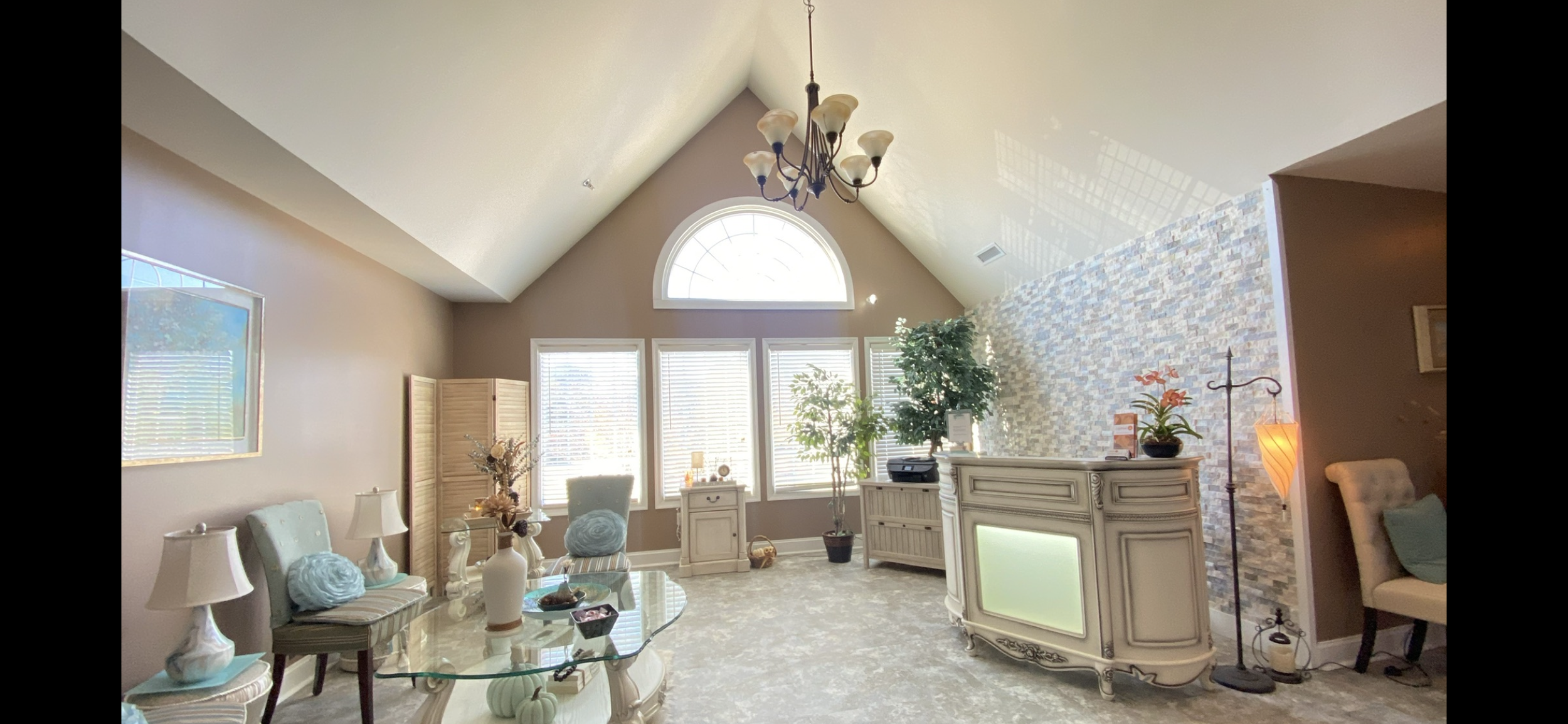 Bright living room with beige walls, vaulted ceiling, chandelier, large arched window, seating area with chairs and pillows, decorative console table, potted plants, and a stone accent wall.