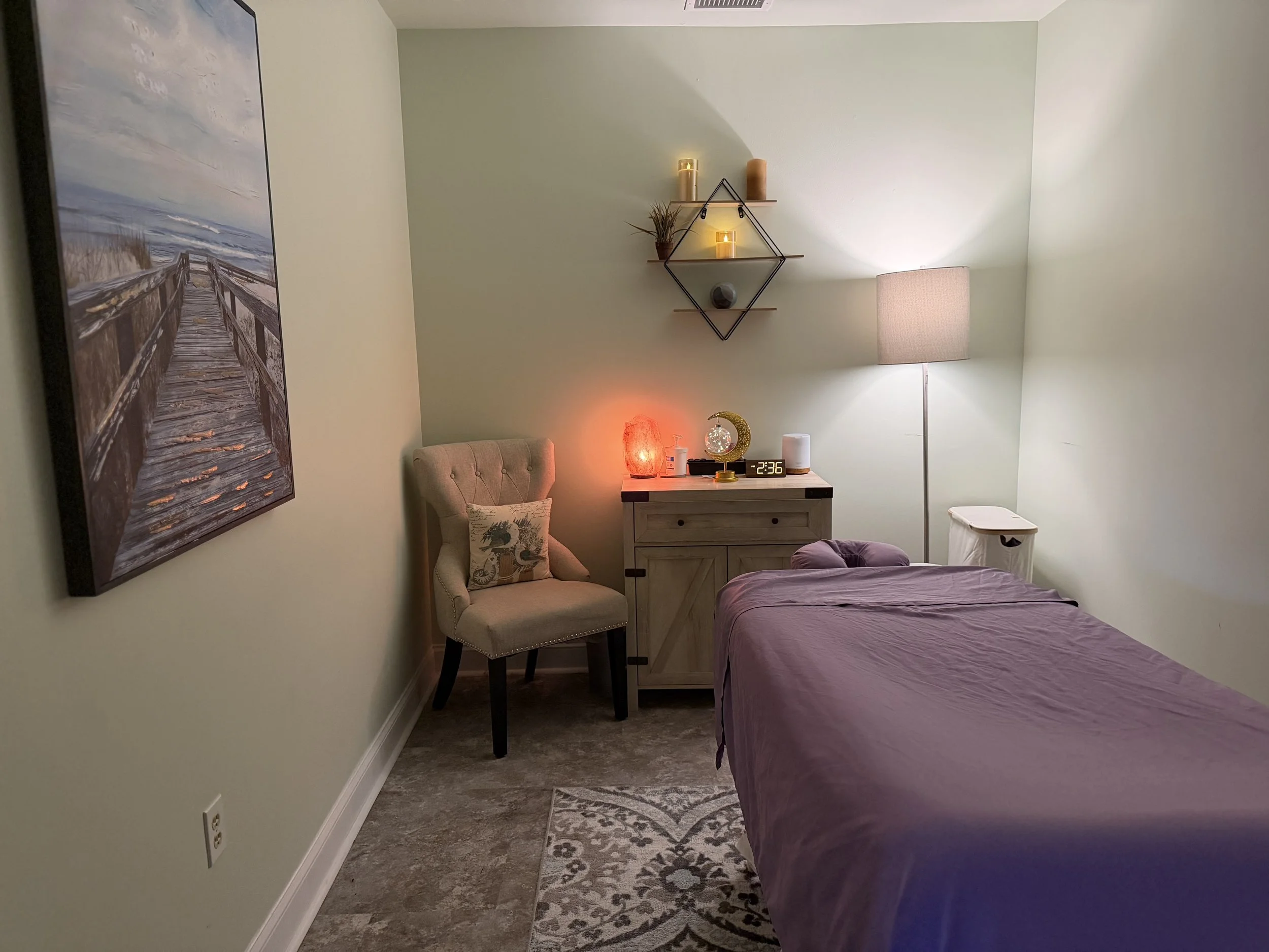 A cozy bedroom with a bed covered in a purple sheet on the right, a beige tufted chair with a decorative pillow on the left, a wooden dresser with a salt lamp, a moon-shaped clock, and a candle, next to a tall floor lamp with a white shade. On the wall above the dresser, there are two wooden shelves with candles and decorative items. A framed picture of a wooden dock and beach scene hangs on the left wall, and a patterned rug is on the floor.