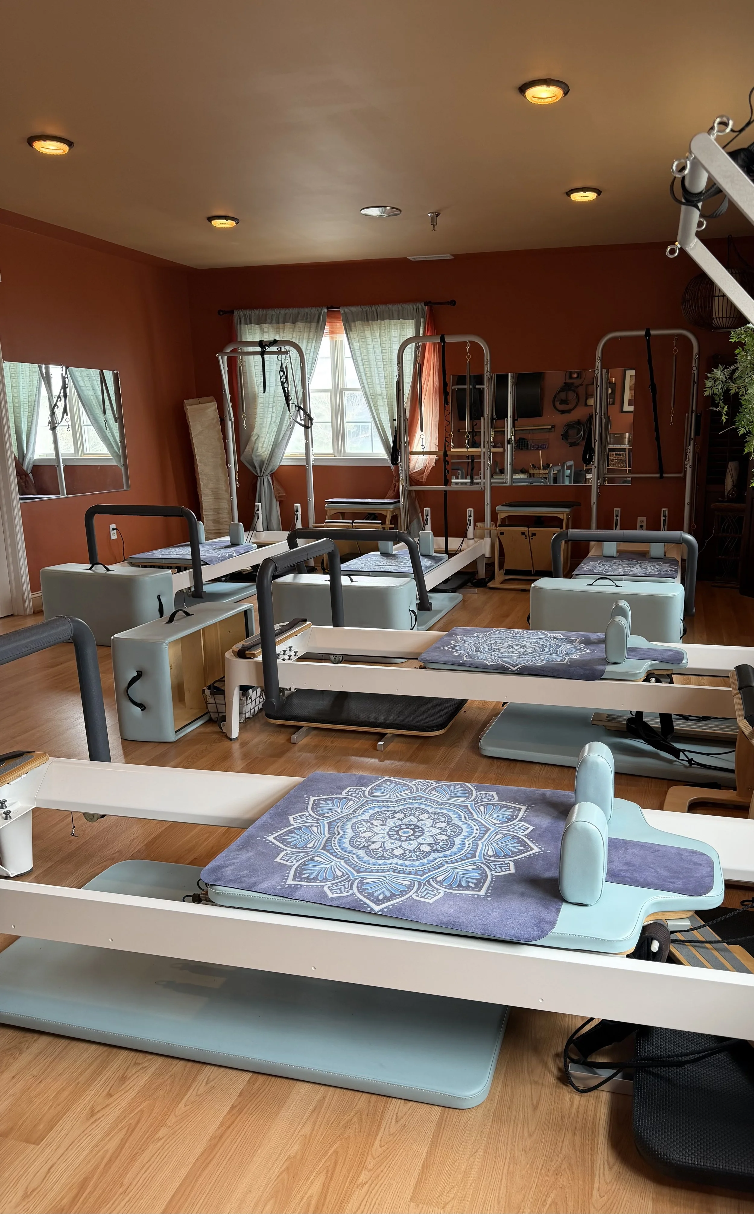 A room set up for Pilates or physical therapy with multiple reformer machines and gym equipment, with natural light coming through windows and curtains.
