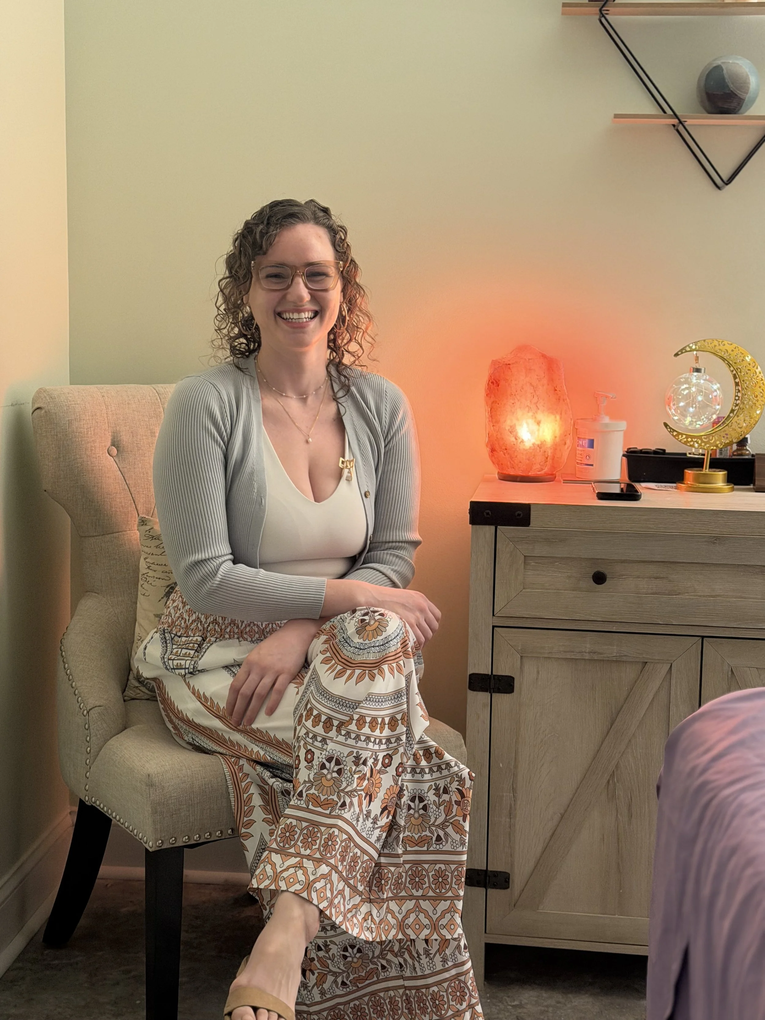 A woman with curly hair, glasses, and a warm smile sitting on a beige upholstered chair in a cozy room, dressed in patterned wide-leg pants and a white top, with a gray cardigan. Behind her is a wooden sideboard with decorative items including a salt lamp, a moon and star ornament, and a pump dispenser, illuminated by soft, warm lighting.