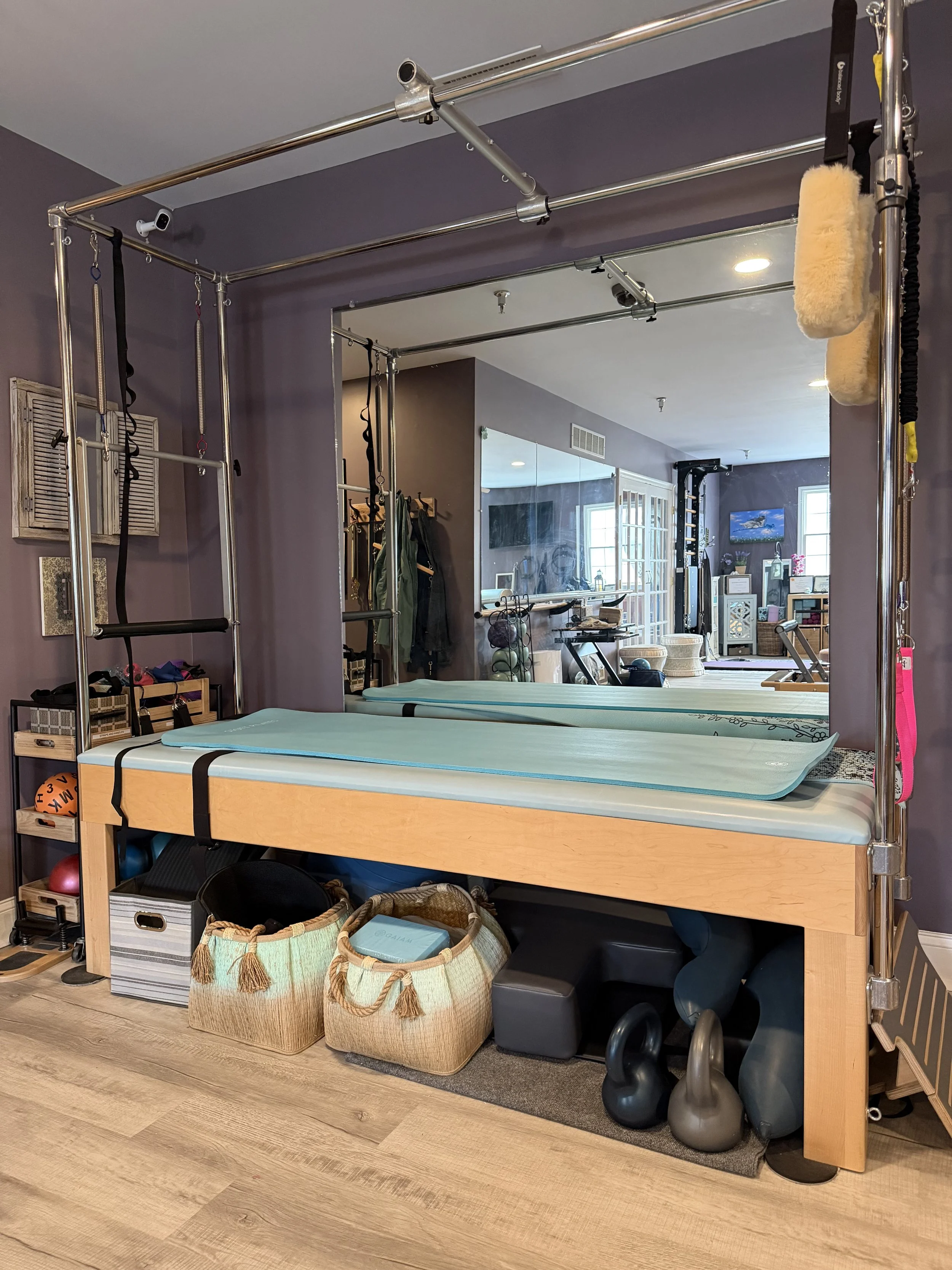 Pilates reformer machine with pads in a fitness room, with kettlebells and baskets underneath, mirror on wall, purple walls, and exercise equipment in background.
