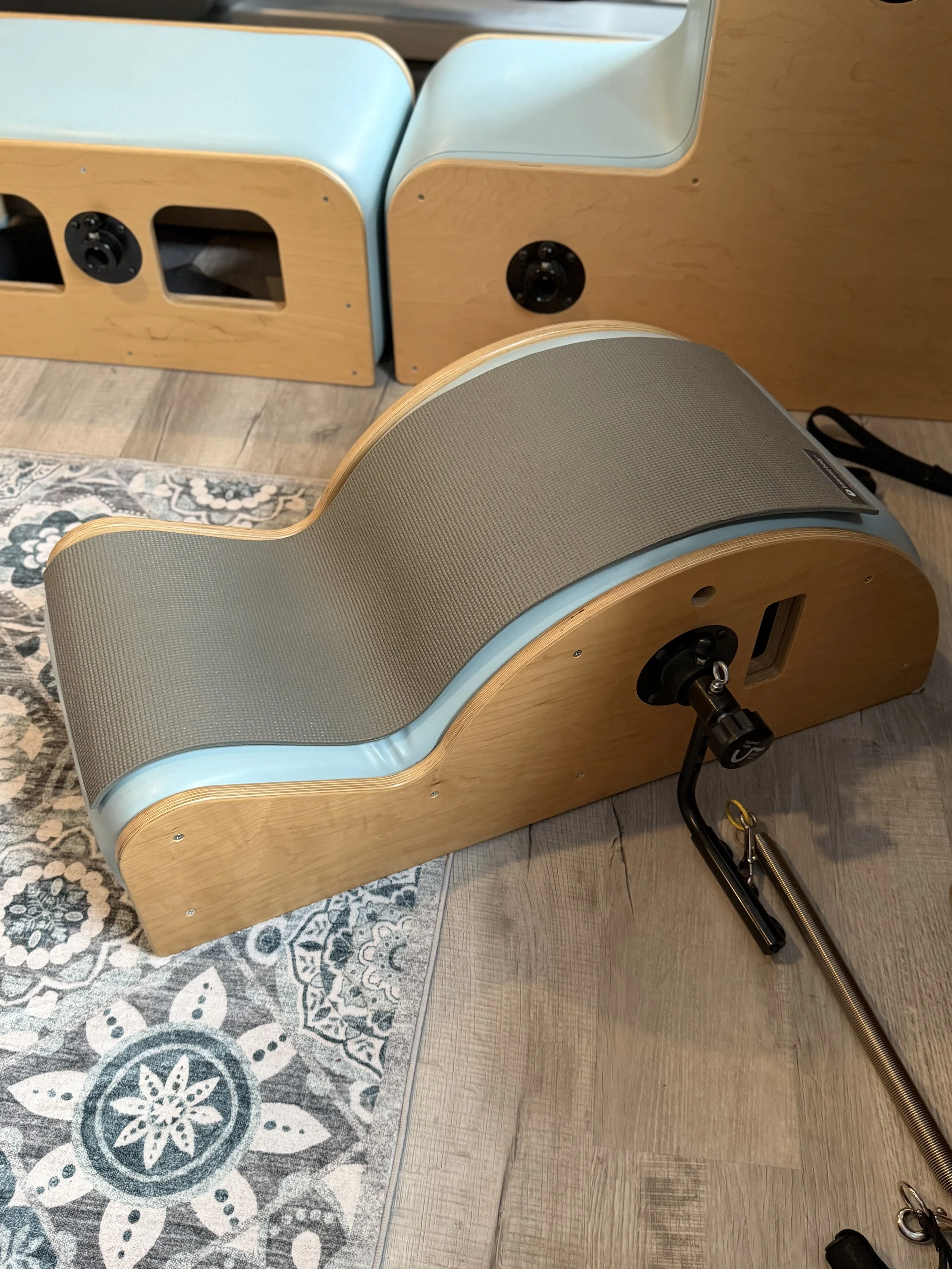 A wooden and fabric device with a curved shape, placed on a wooden floor next to a patterned rug, with a black clamp attached to its side.