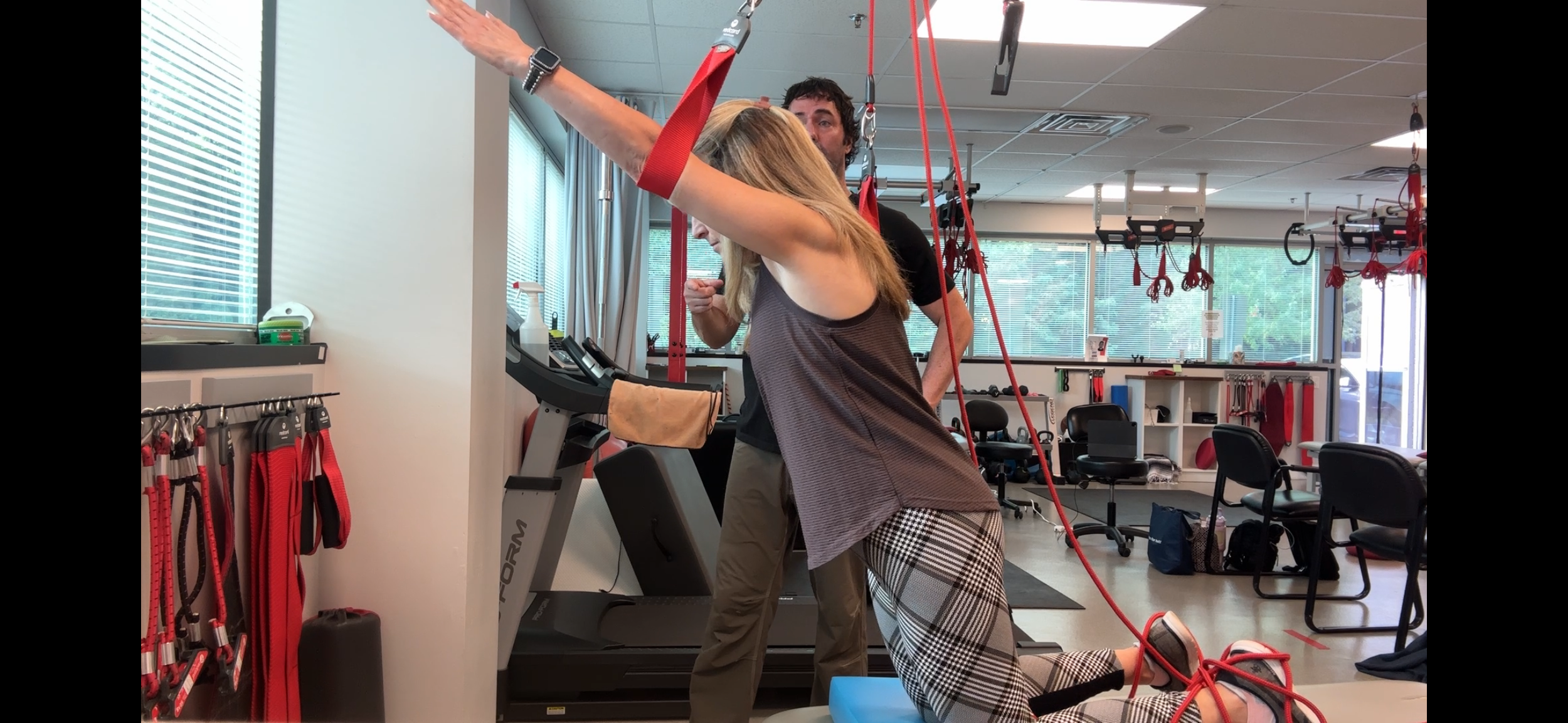 A woman doing a kneeling exercise on a blue mat with resistance bands attached to a wall, with a trainer guiding her in a gym or physical therapy center.