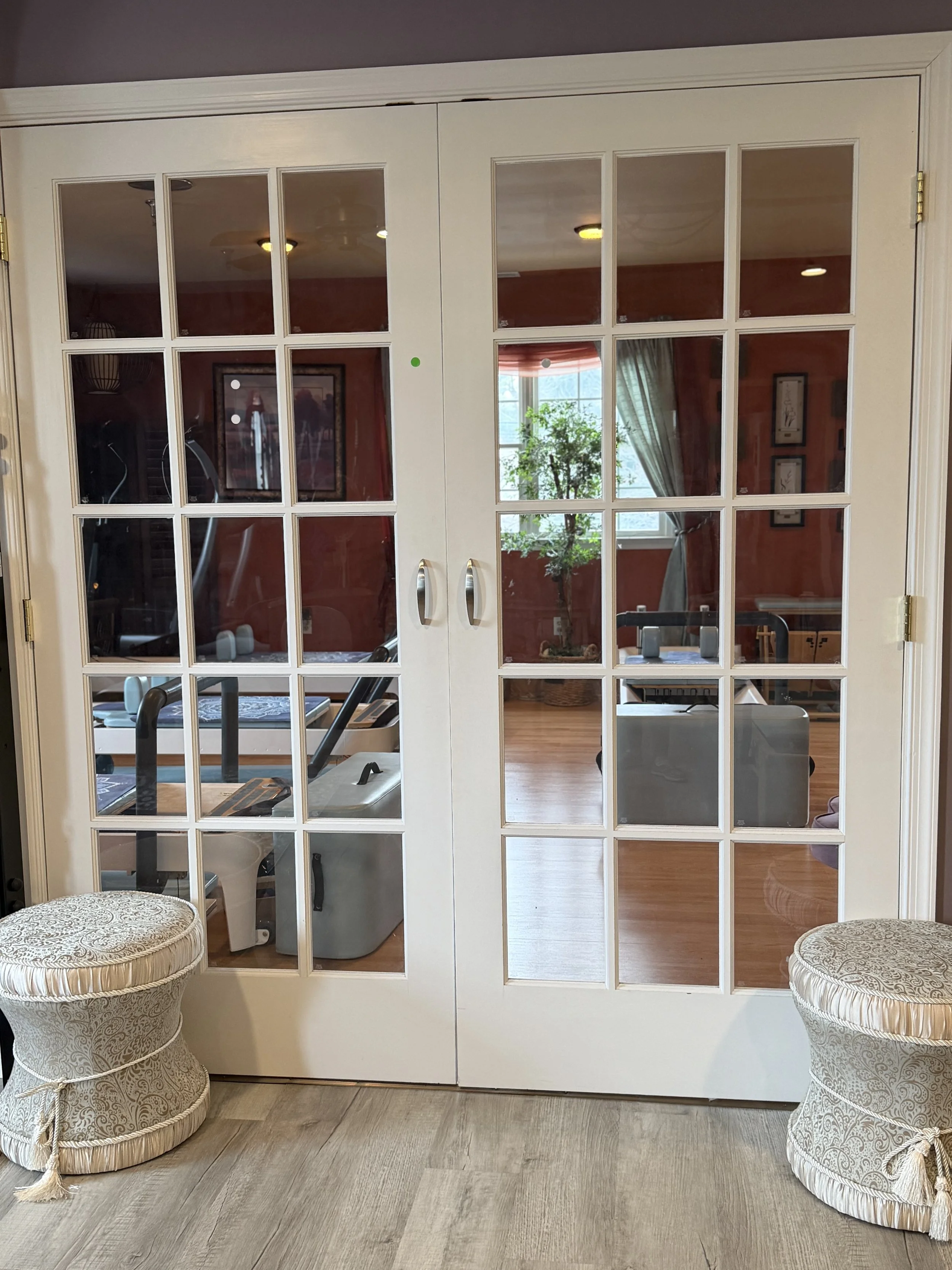 A set of white French doors with glass panels leading into a room with gym equipment, a window with curtains, and a potted plant. Two decorative poufs are placed on either side of the doors on a wood floor.