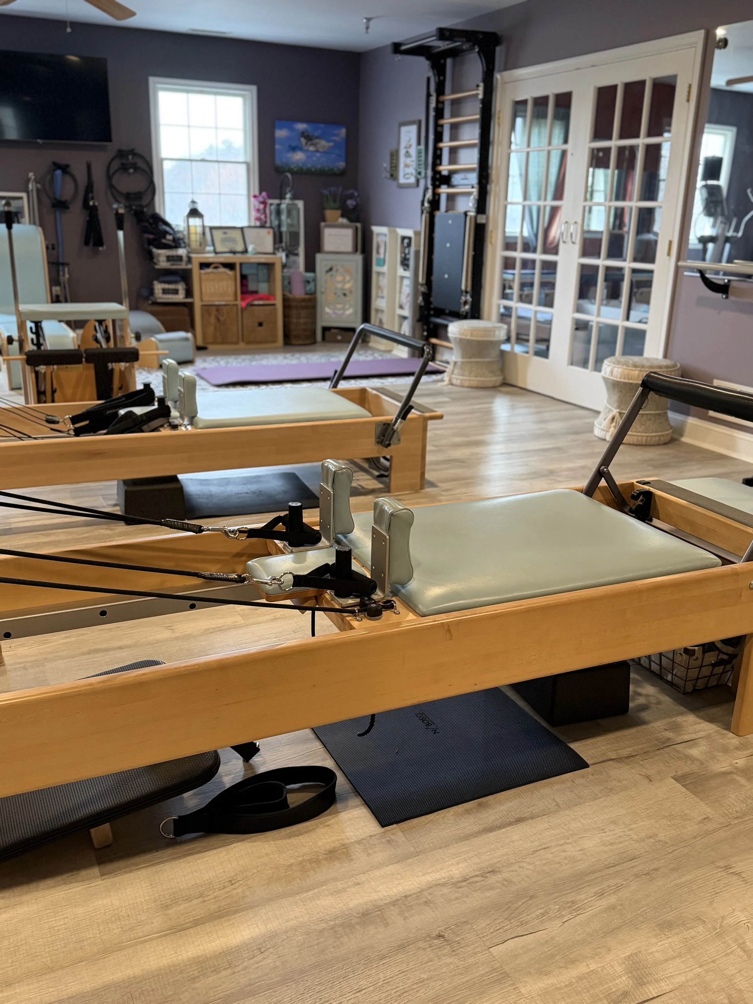 Indoor Pilates or physical therapy studio with reformer machines, a yoga mat, and various exercise equipment and storage units in the background.