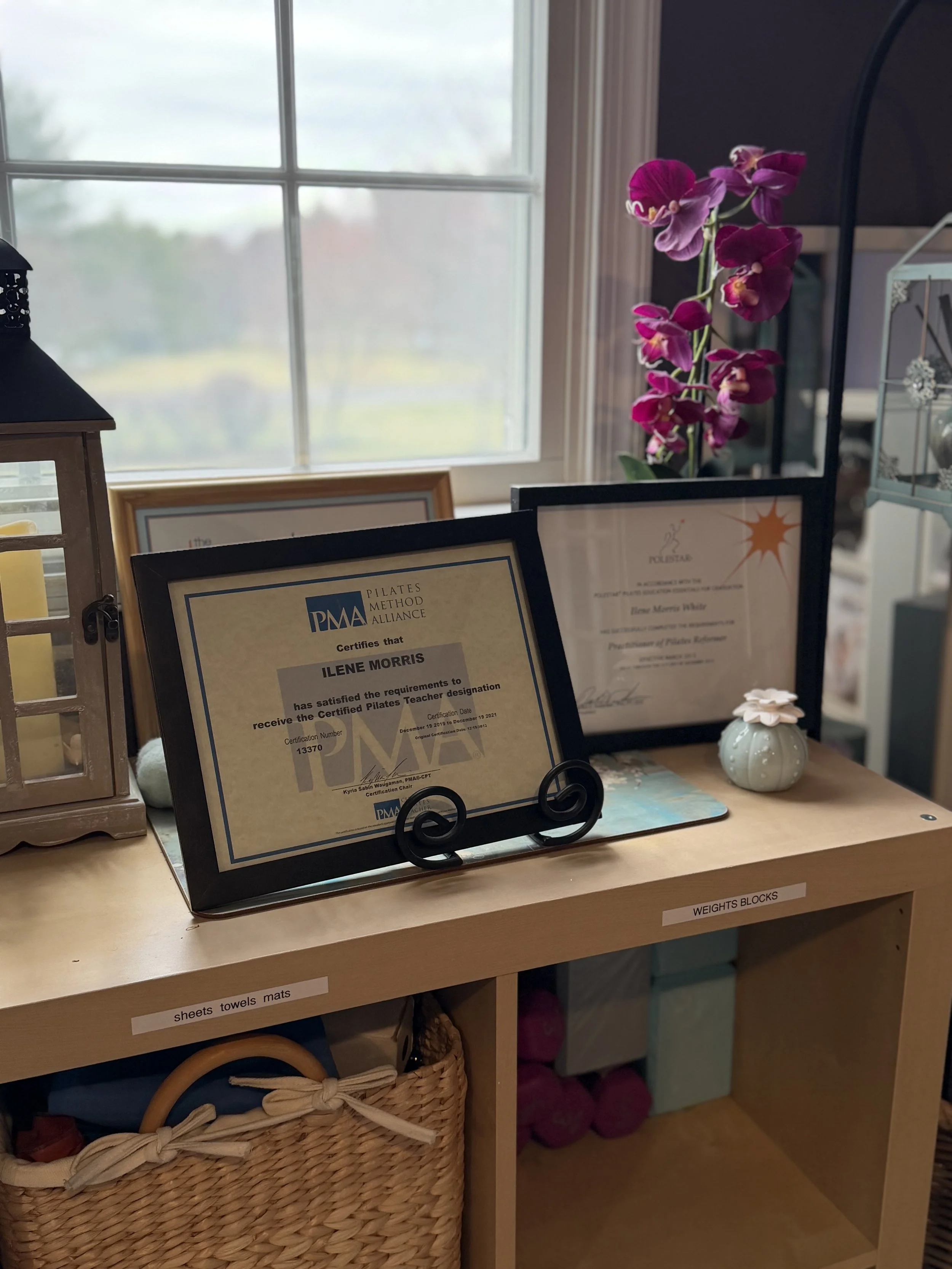 Certificates on a black stand, a purple orchid, a small ceramic pumpkin, and a glass lantern with a candle inside on a wooden shelf near a window.