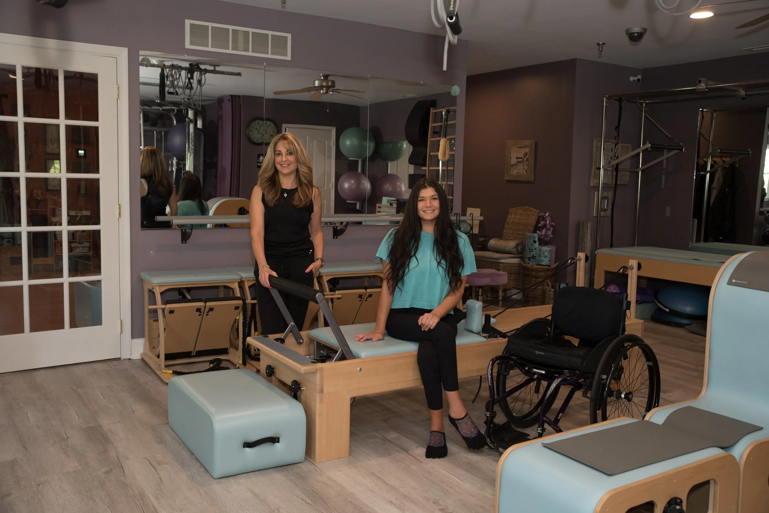 Two women in a physical therapy or rehabilitation center, one sitting on a therapy table and the other standing beside her, with wheelchair and therapy equipment in the background.