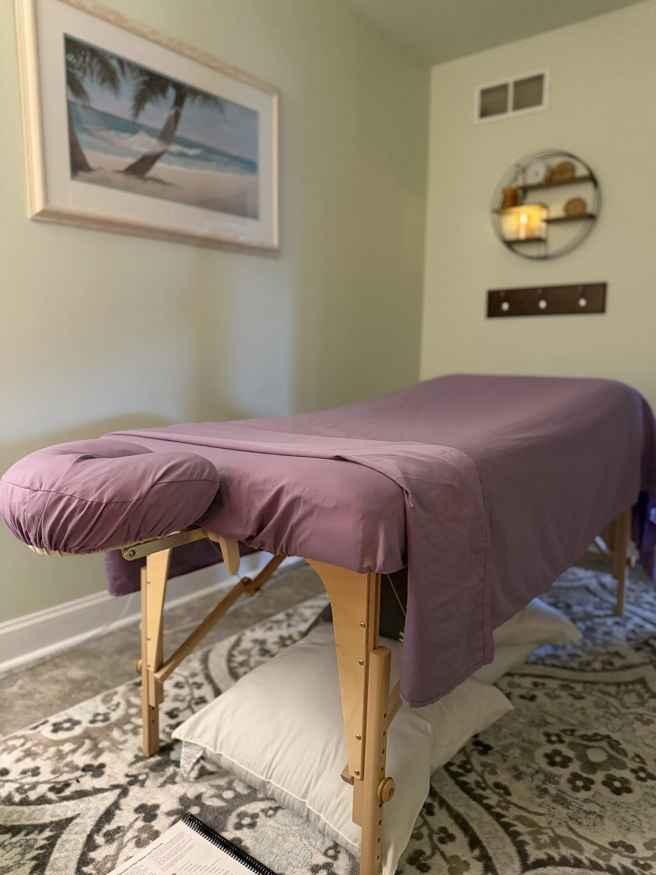 A massage table with a purple sheet in a decorated room, with a pillow underneath at the head, on a patterned rug.