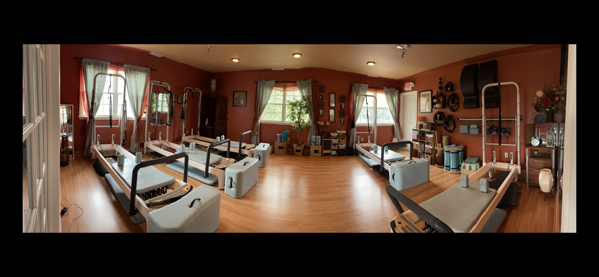 A spacious Pilates studio with wood flooring and burnt orange walls, equipped with multiple Pilates reformer machines, yoga props, and organized shelves with decorative items and plants, illuminated by large windows with curtains.