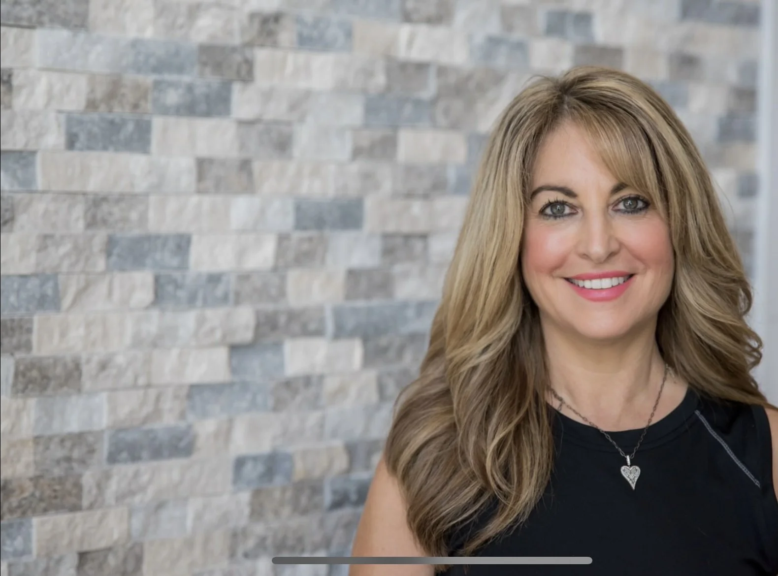 A woman with long wavy blonde hair smiling, wearing a black sleeveless top and a silver heart necklace, standing in front of a gray brick wall.