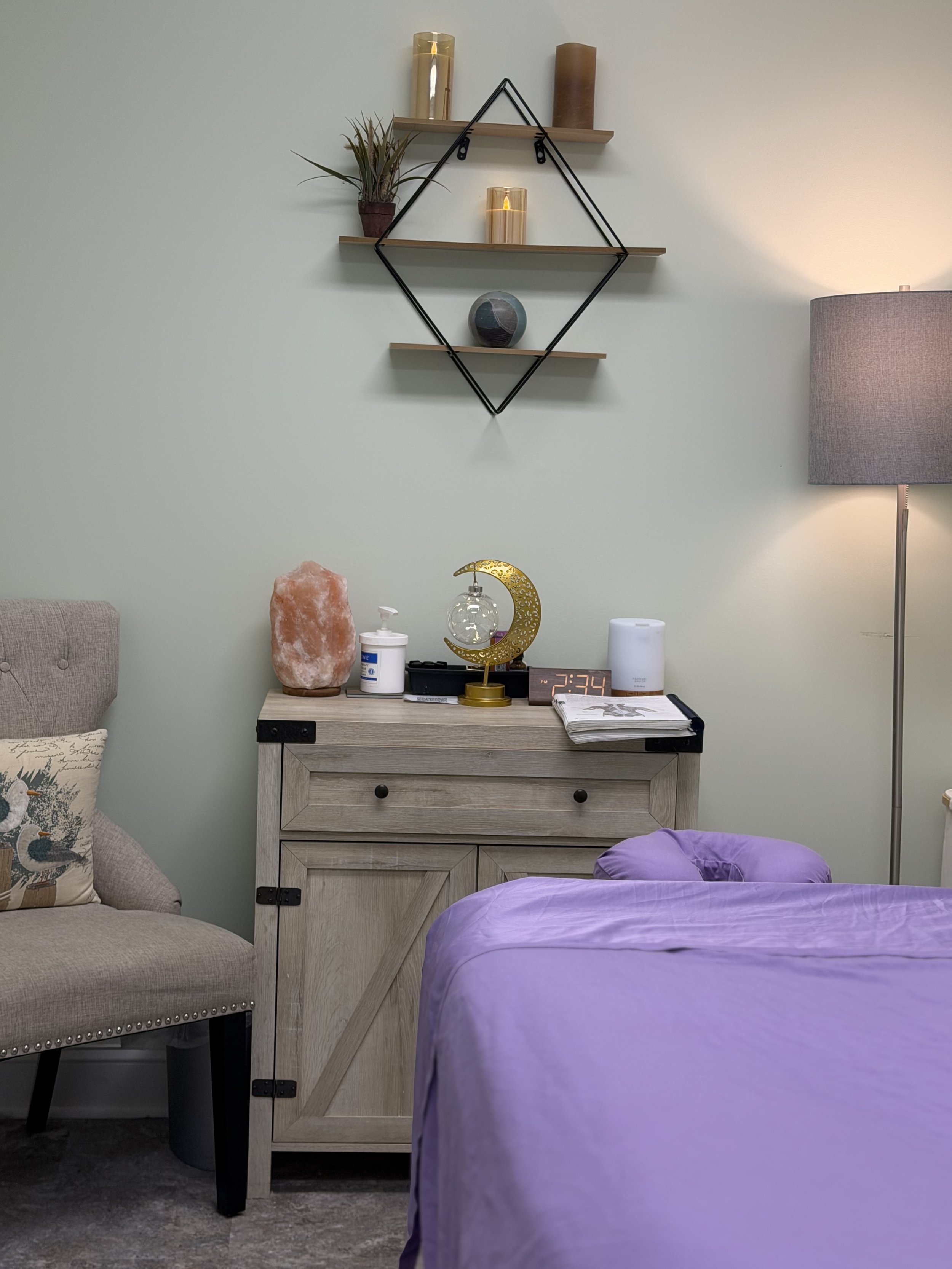 A bedroom corner featuring a bed with purple bedding, a beige armchair with a decorative pillow, a wooden nightstand with a salt lamp, moon-shaped light, a digital clock, and various items, with a green wall and wall shelves decorated with candles, a plant, and a decorative sphere.