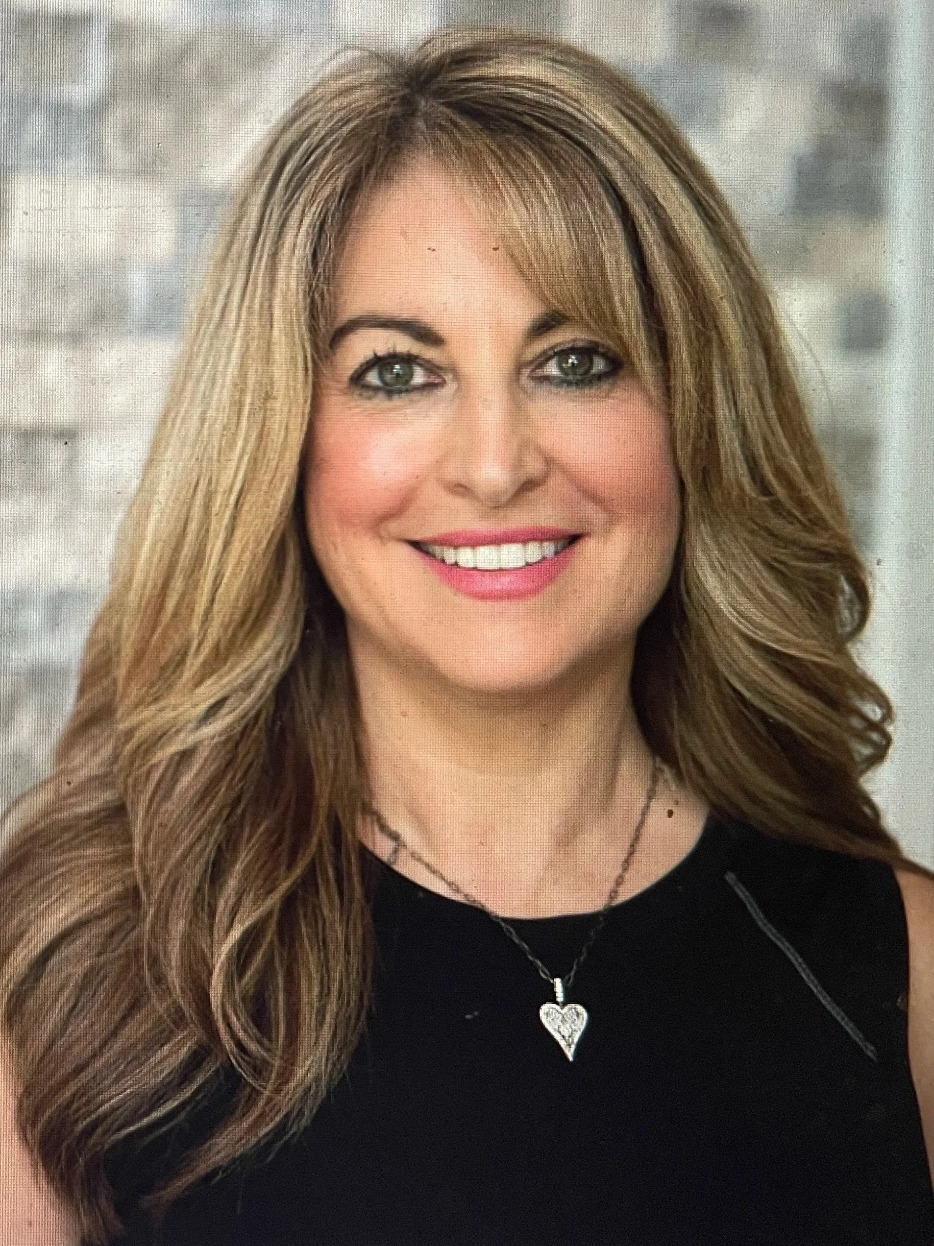 A photo of a woman with long, wavy blonde hair, smiling, wearing a black top, a silver heart necklace, and makeup, against a blurred gray brick wall background.