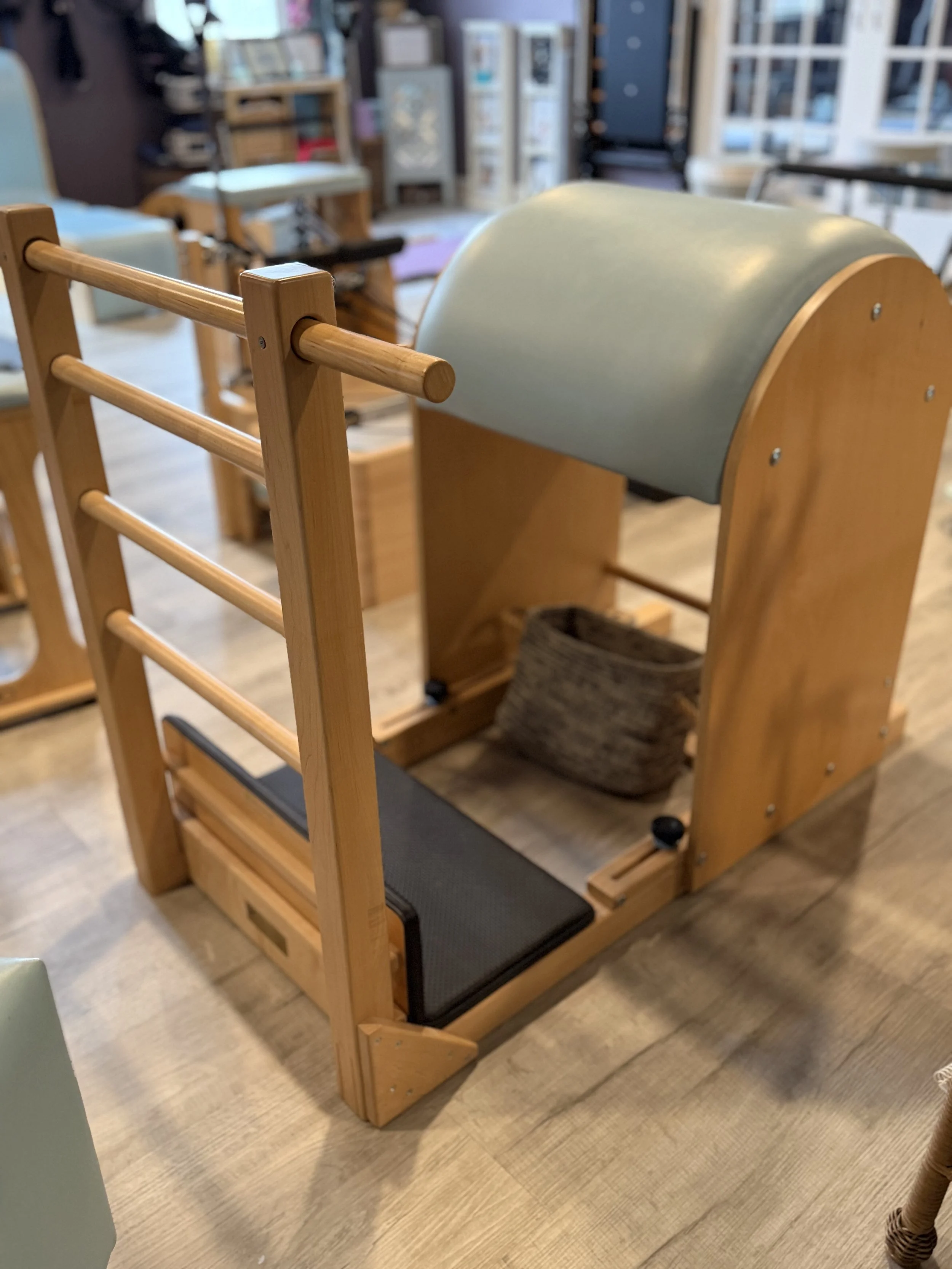 A wooden foot and leg exercise machine with a padded roller and a black cushioned seat in a store or showroom setting.