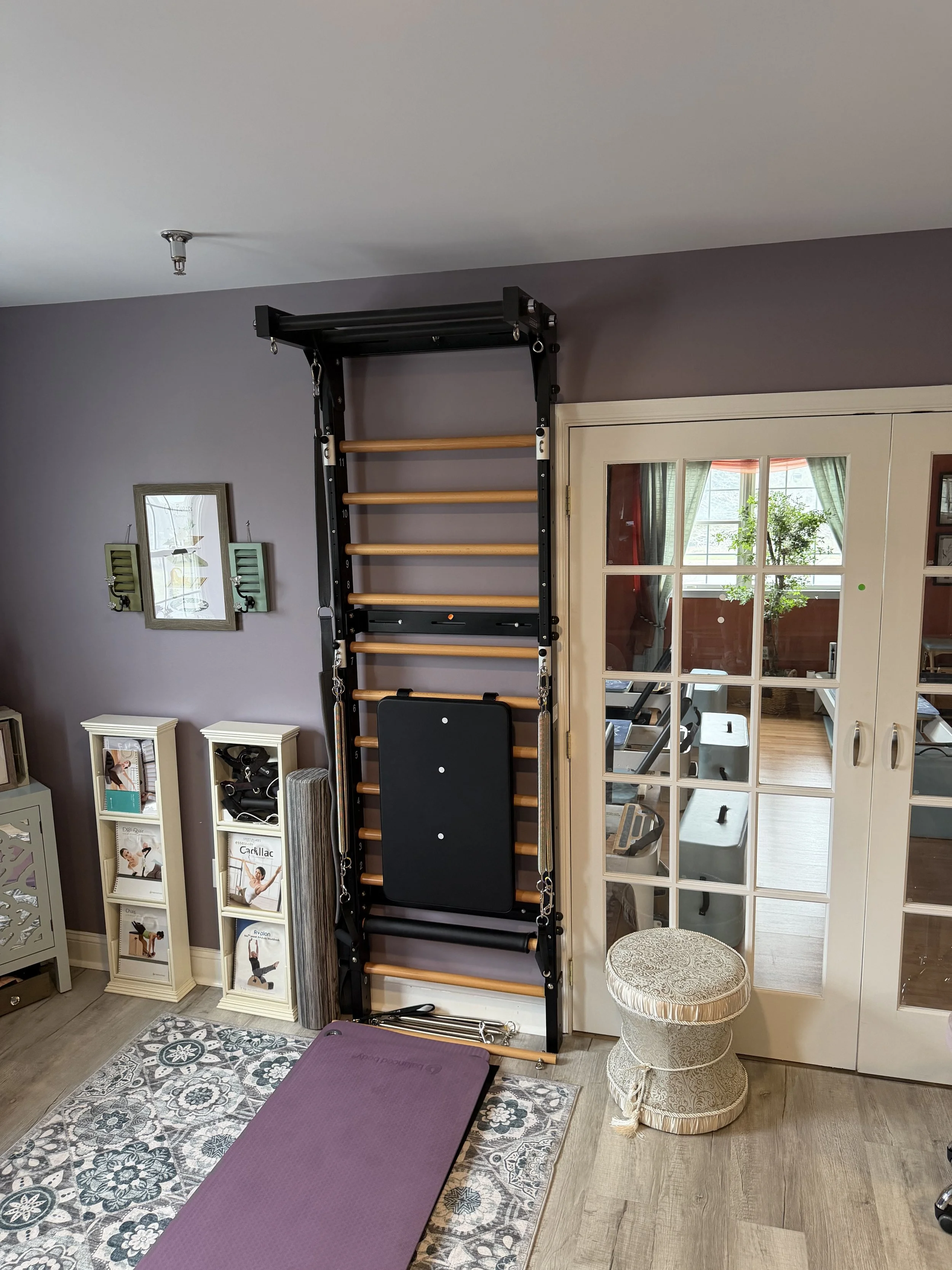 Home gym wall with a black and wood exercise frame and a purple yoga mat on a patterned rug, next to slatted storage units and a decorative pouf, inside a room with a door leading to a sunlit space with green curtains and potted plant.