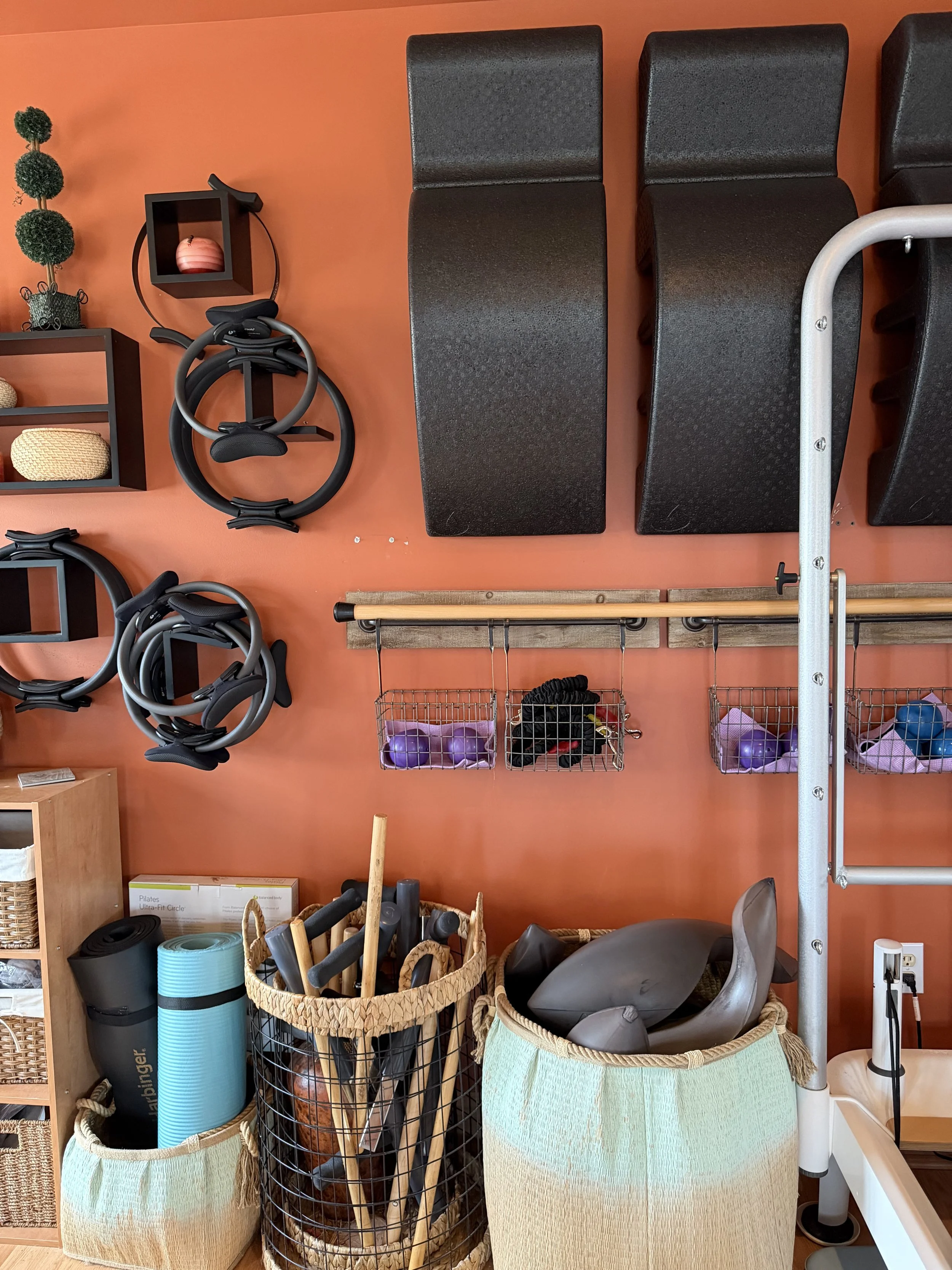 Fitness studio wall with black workout benches, exercise rings, and wall-mounted baskets containing small exercise balls and gloves. Below are baskets holding yoga mats, weights, and other workout gear.