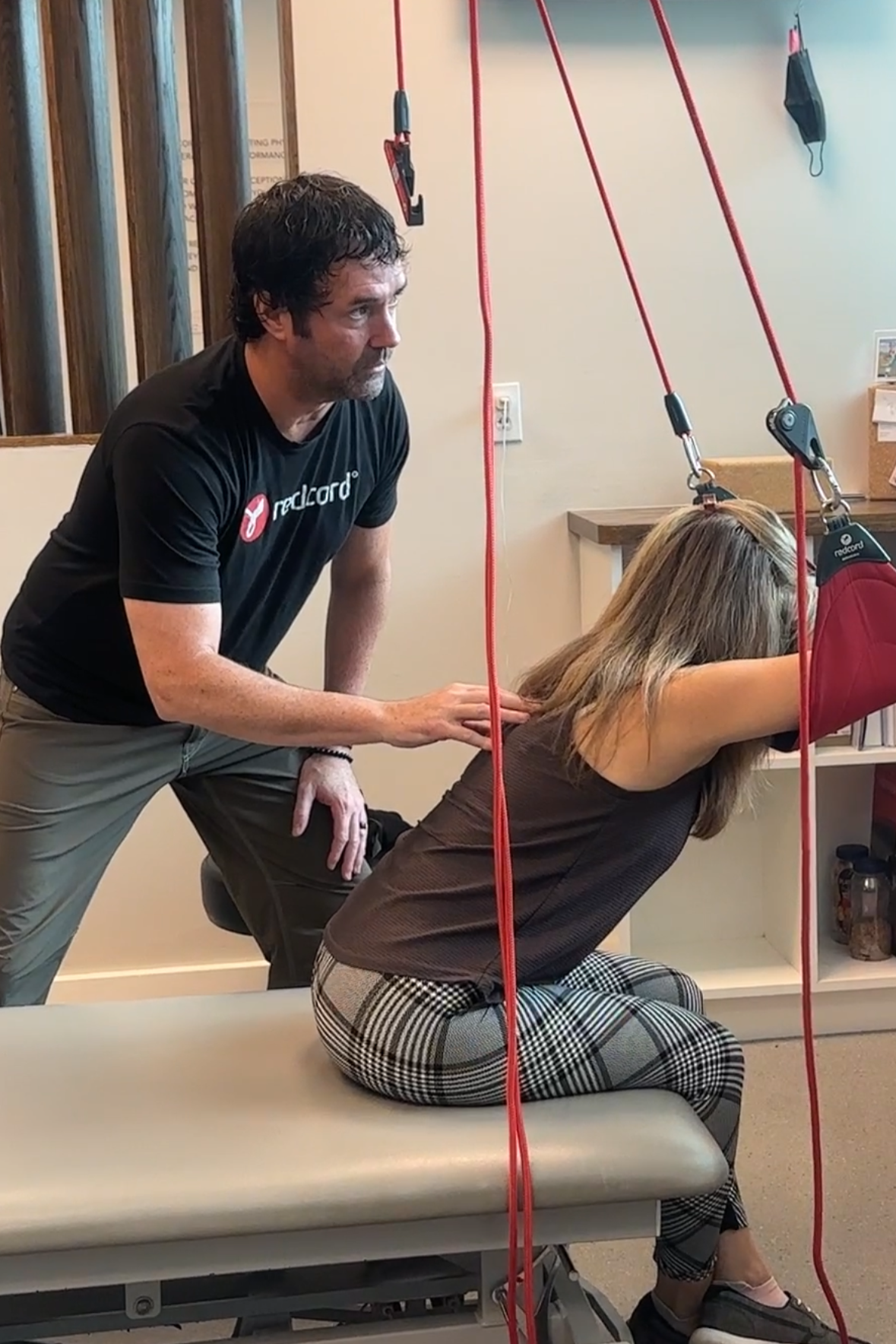 Medical professional performing a therapy on a woman sitting on an examination table, with red straps hanging from the ceiling.
