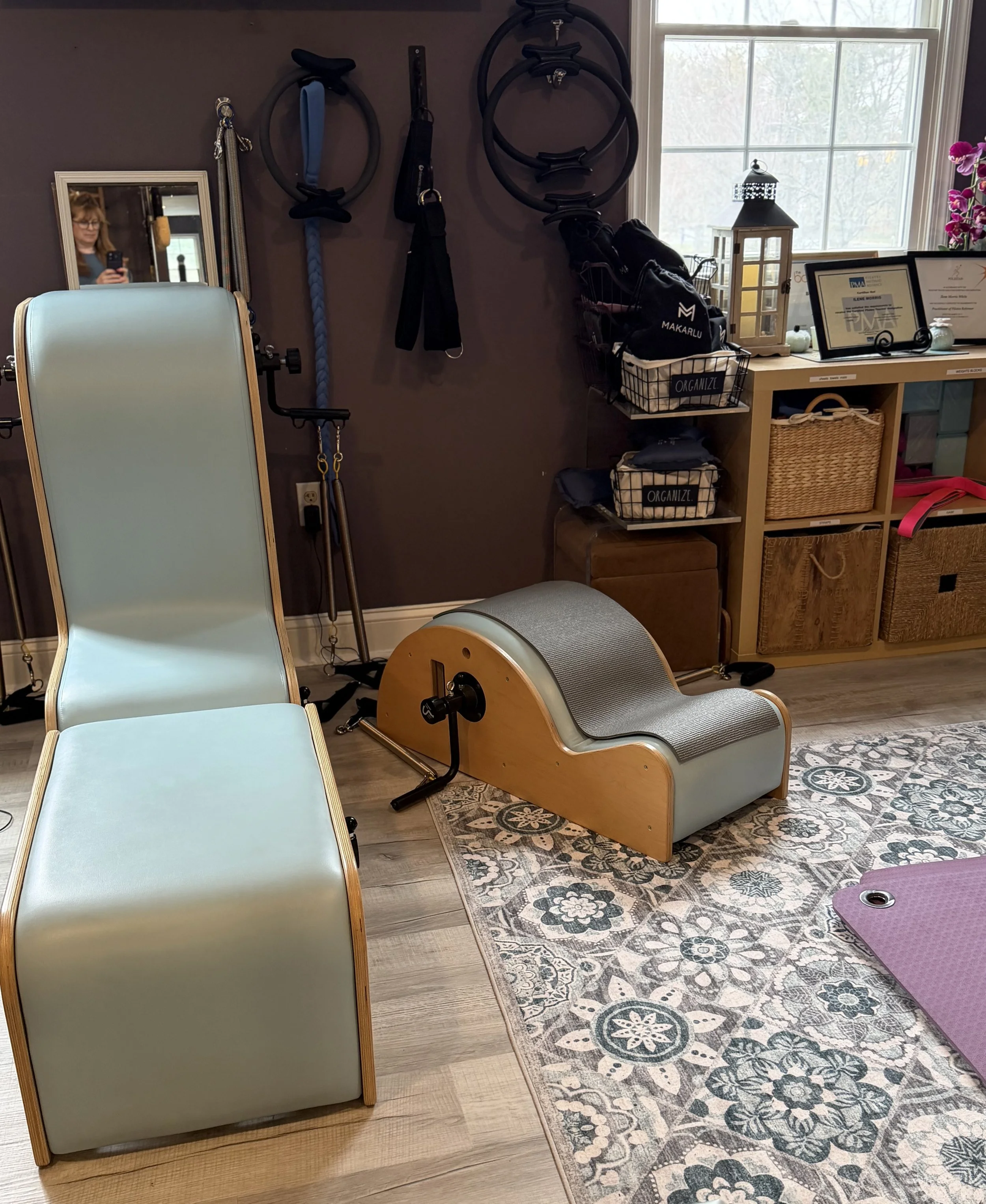 Interior of a Pilates studio with a reformer machine, a chair, wall-mounted resistance rings, and a window with a decorative jar and framed certificates on a shelf.