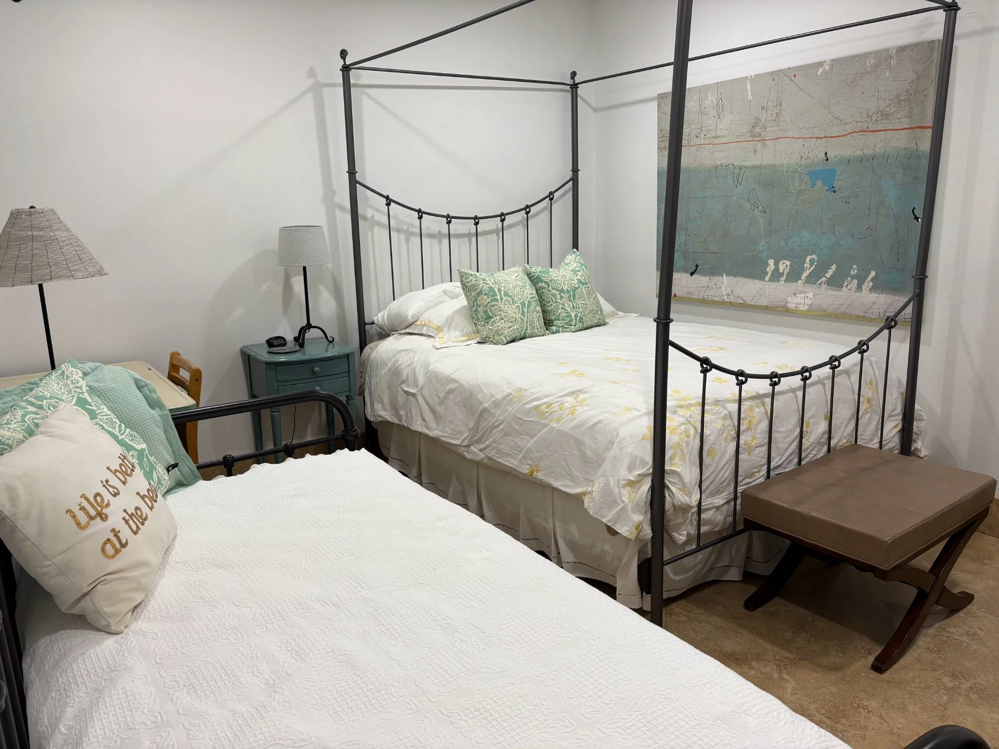 Two beds in a bedroom with decorative pillows, a nightstand with a lamp and alarm clock, a bedside table with a lamp, and a piece of artwork on the wall behind one bed.