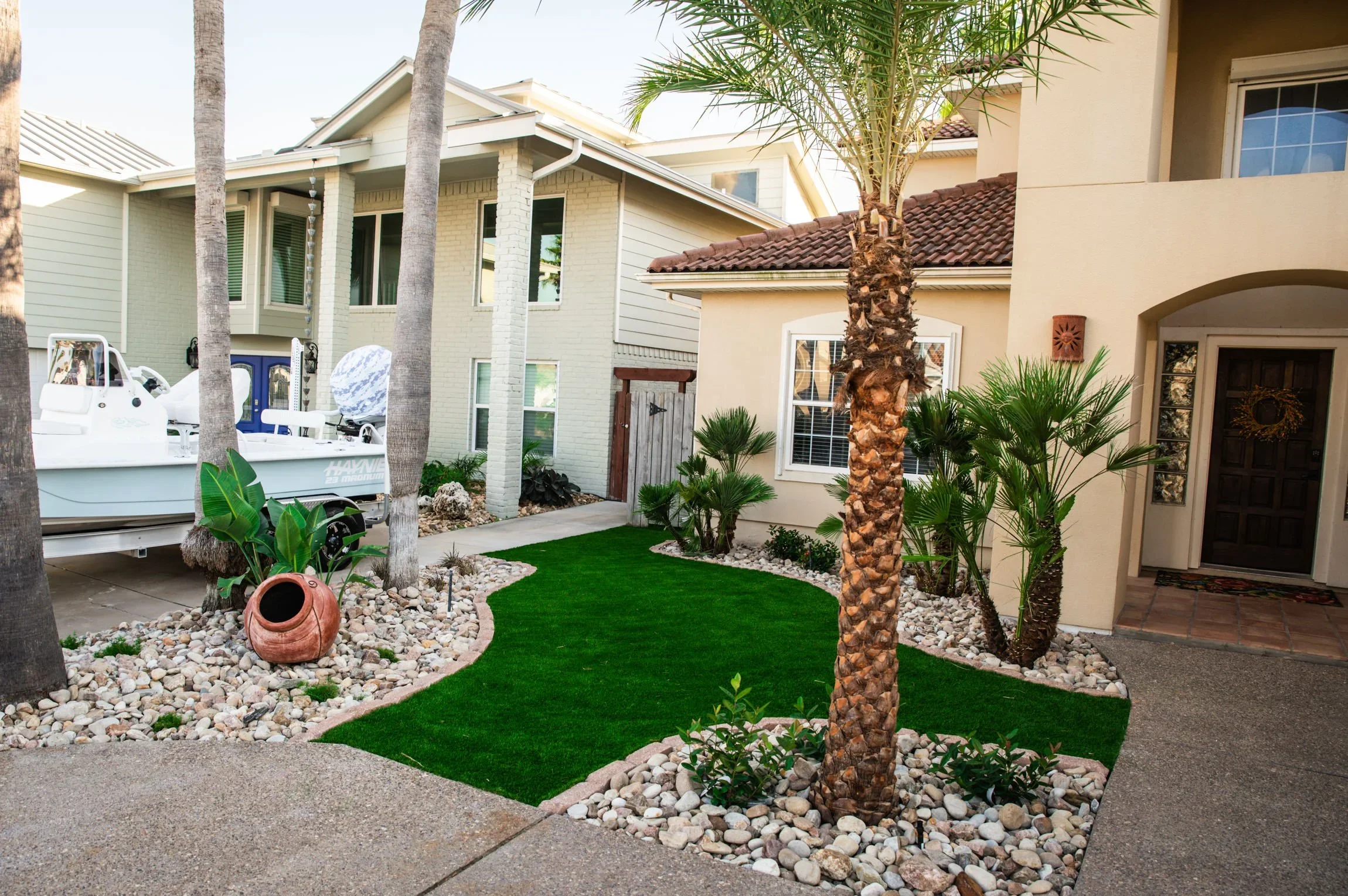 Front yard landscaping with palm trees, green grass, small shrubs, white and beige houses, a boat on a trailer, and decorative rocks.