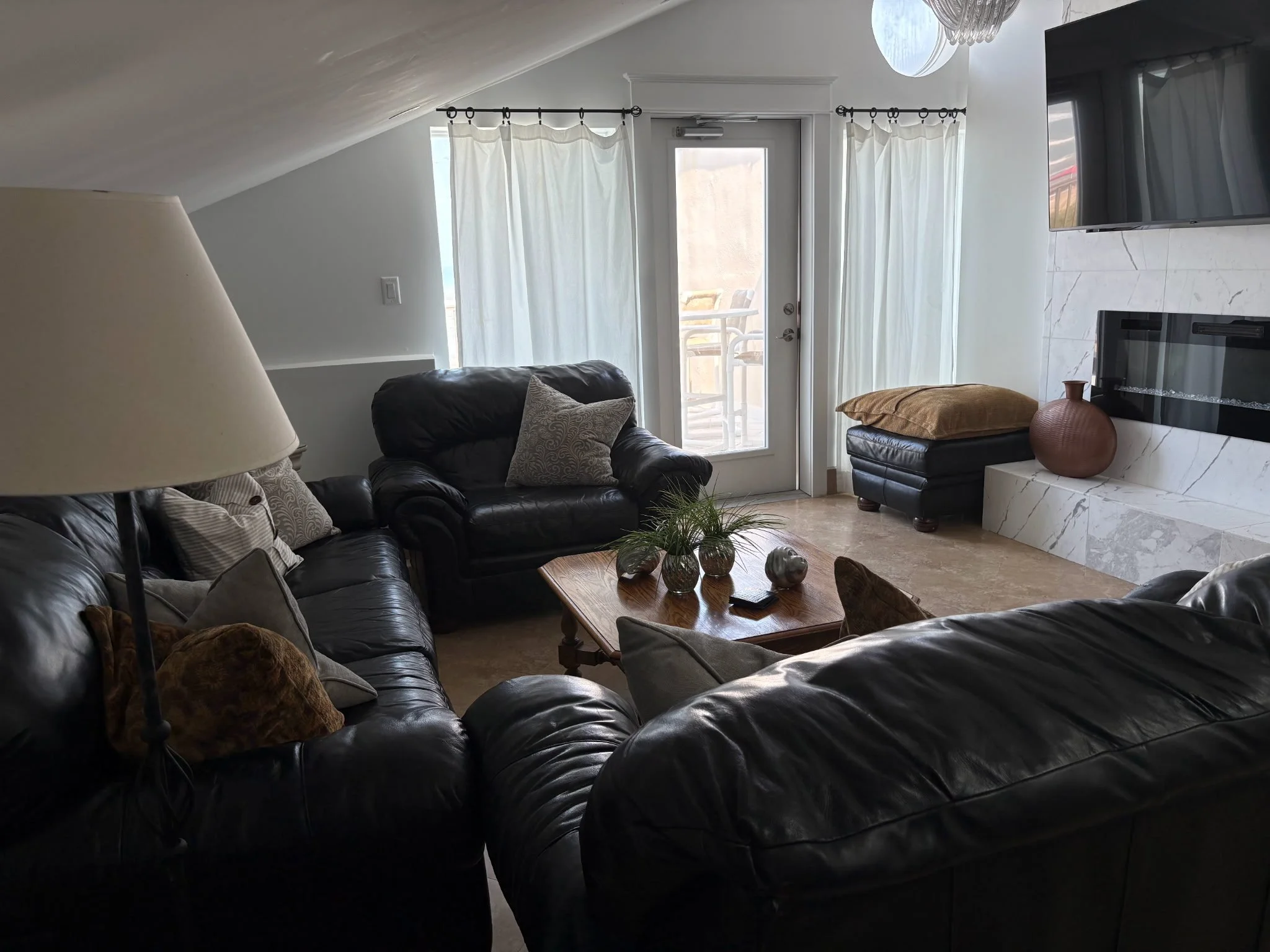 Living room with black leather sofas, a wooden coffee table, white curtains, a door leading outside, a wall-mounted TV, and a fireplace.