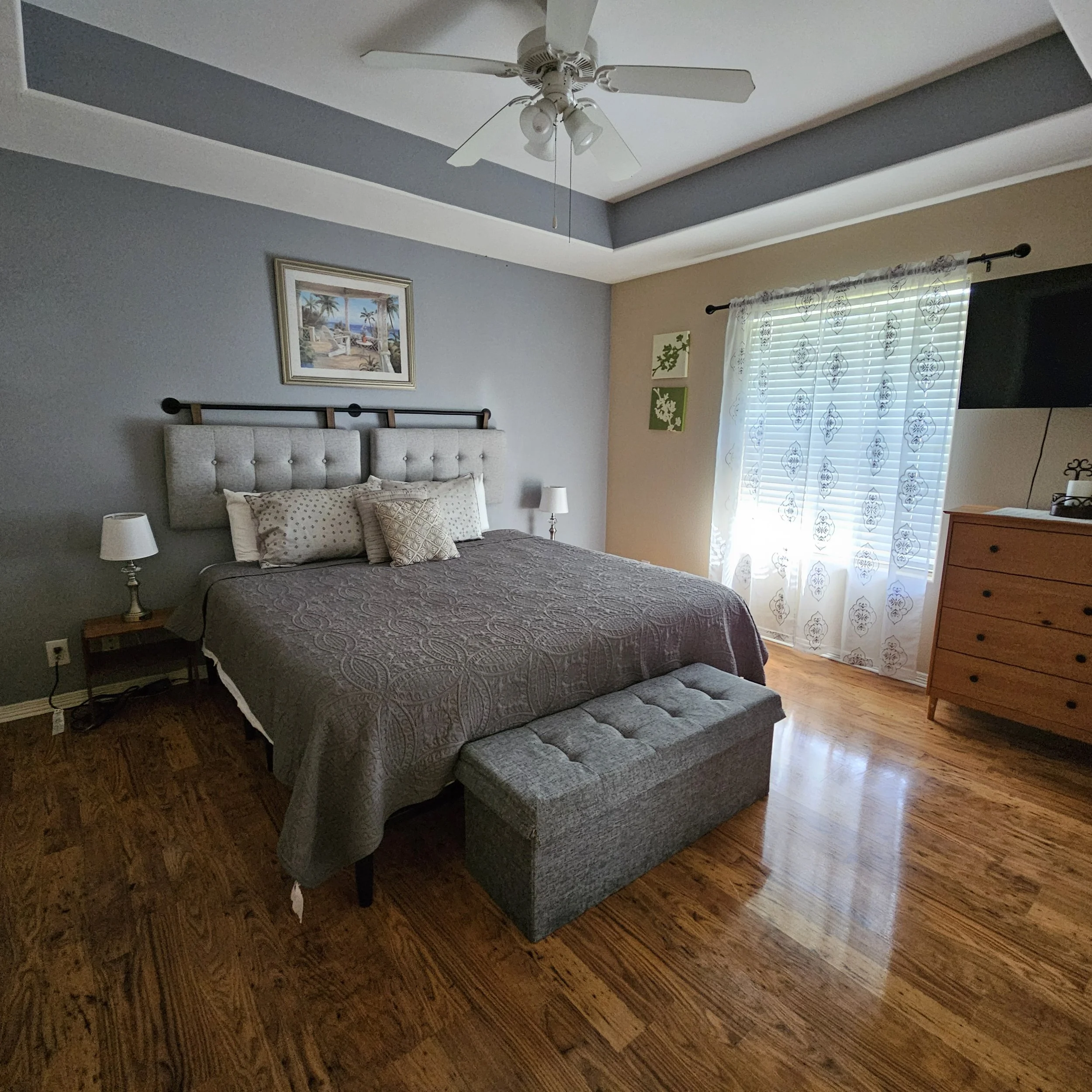 A bedroom with a gray accent wall, wooden floor, a bed with tufted gray headboard, gray and beige bedding, two side tables with lamps, a wooden dresser, a window with patterned curtains, framed artwork, and a ceiling fan.